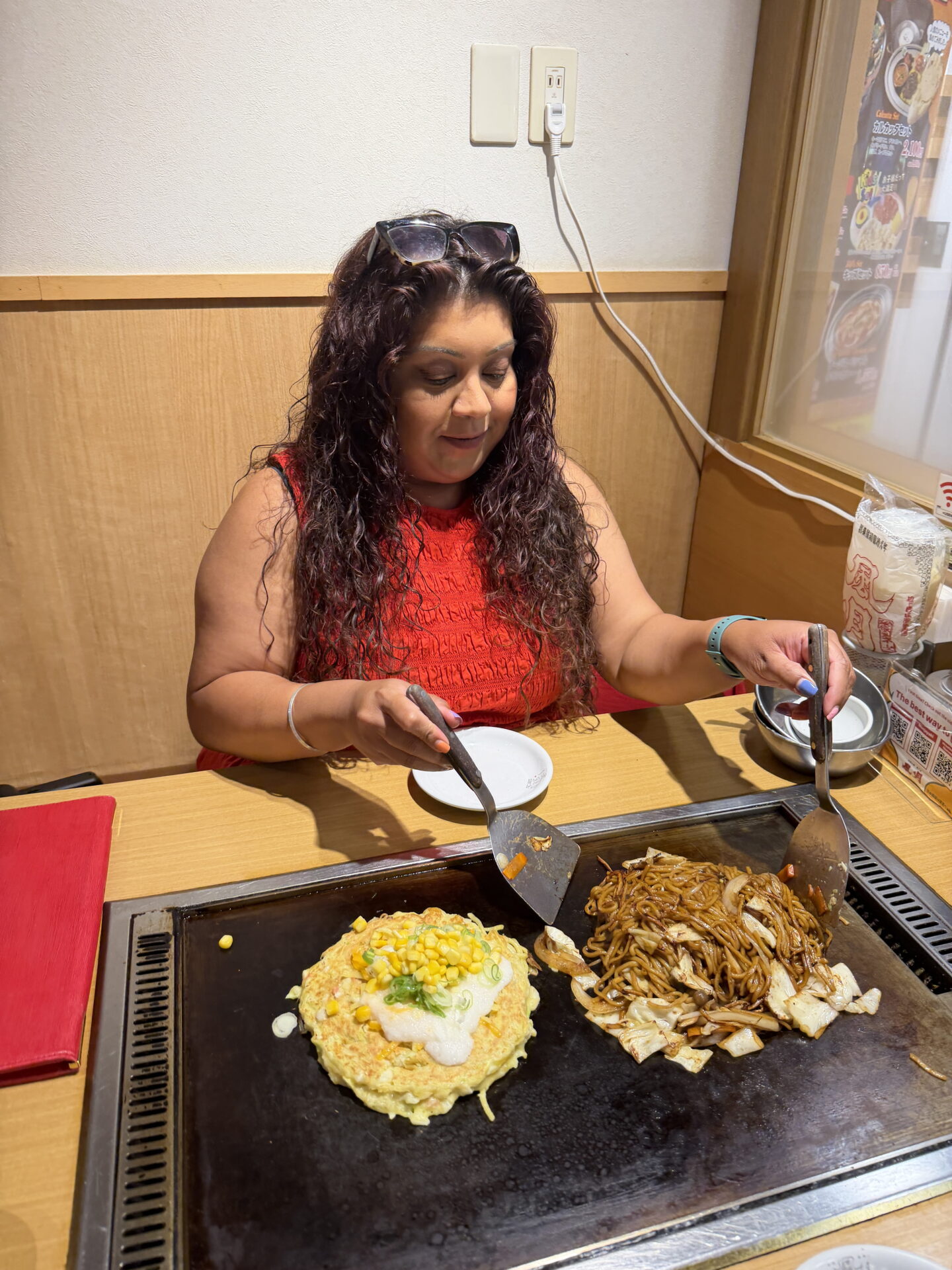 Nishi in a bright red sleeveless top uses two metal spatulas to cook Japanese yakisoba noodles with cabbage alongside okonomiyaki topped with sweetcorn and spring onions. Both dishes are cooking on a large hotplate built into the restaurant table.