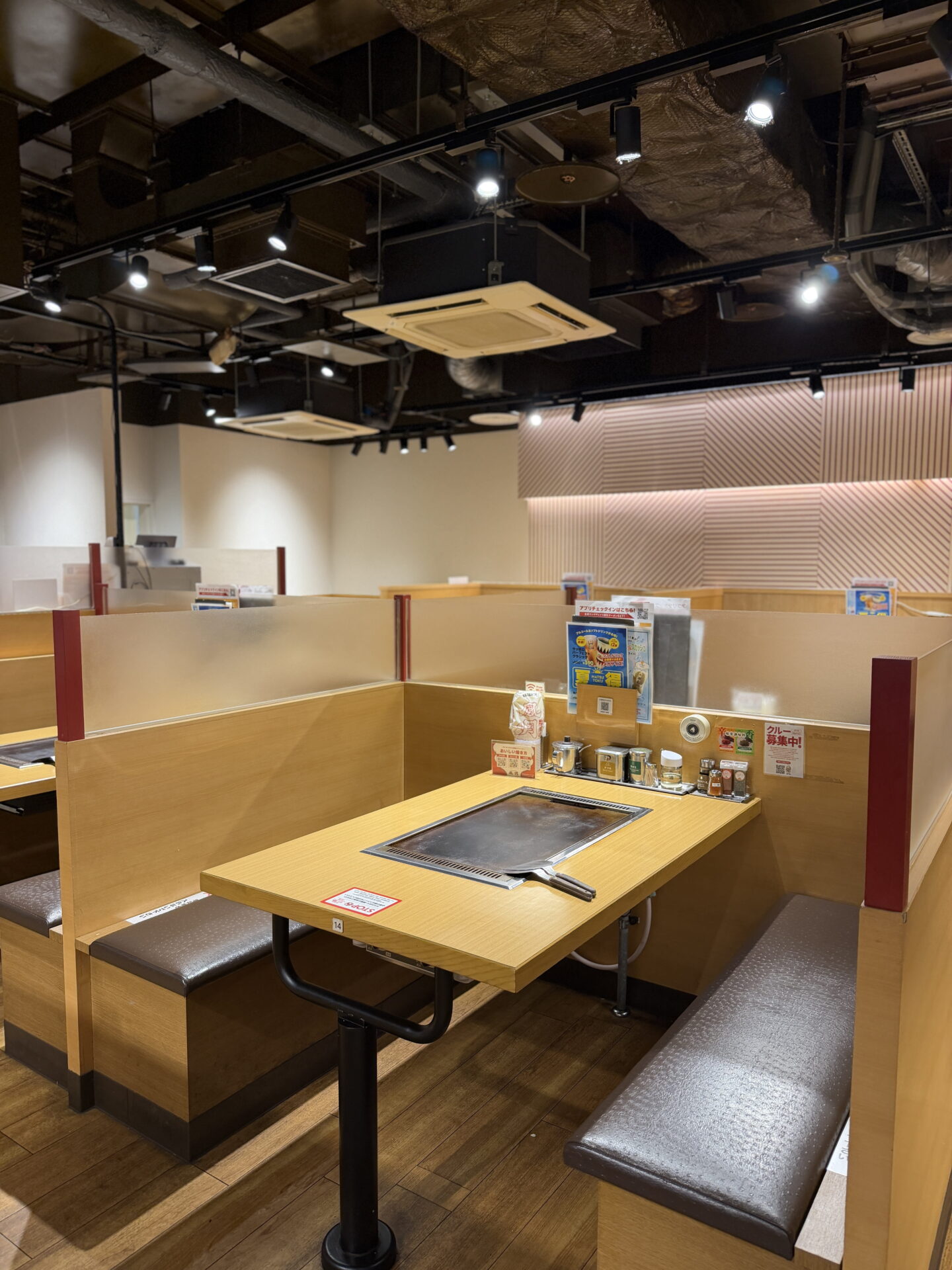 An empty Japanese restaurant with wooden booth seating, frosted glass partitions and built in teppan hotplates at each table. Condiments and promotional signs sit along the tables beneath exposed black ceilings and spotlights.