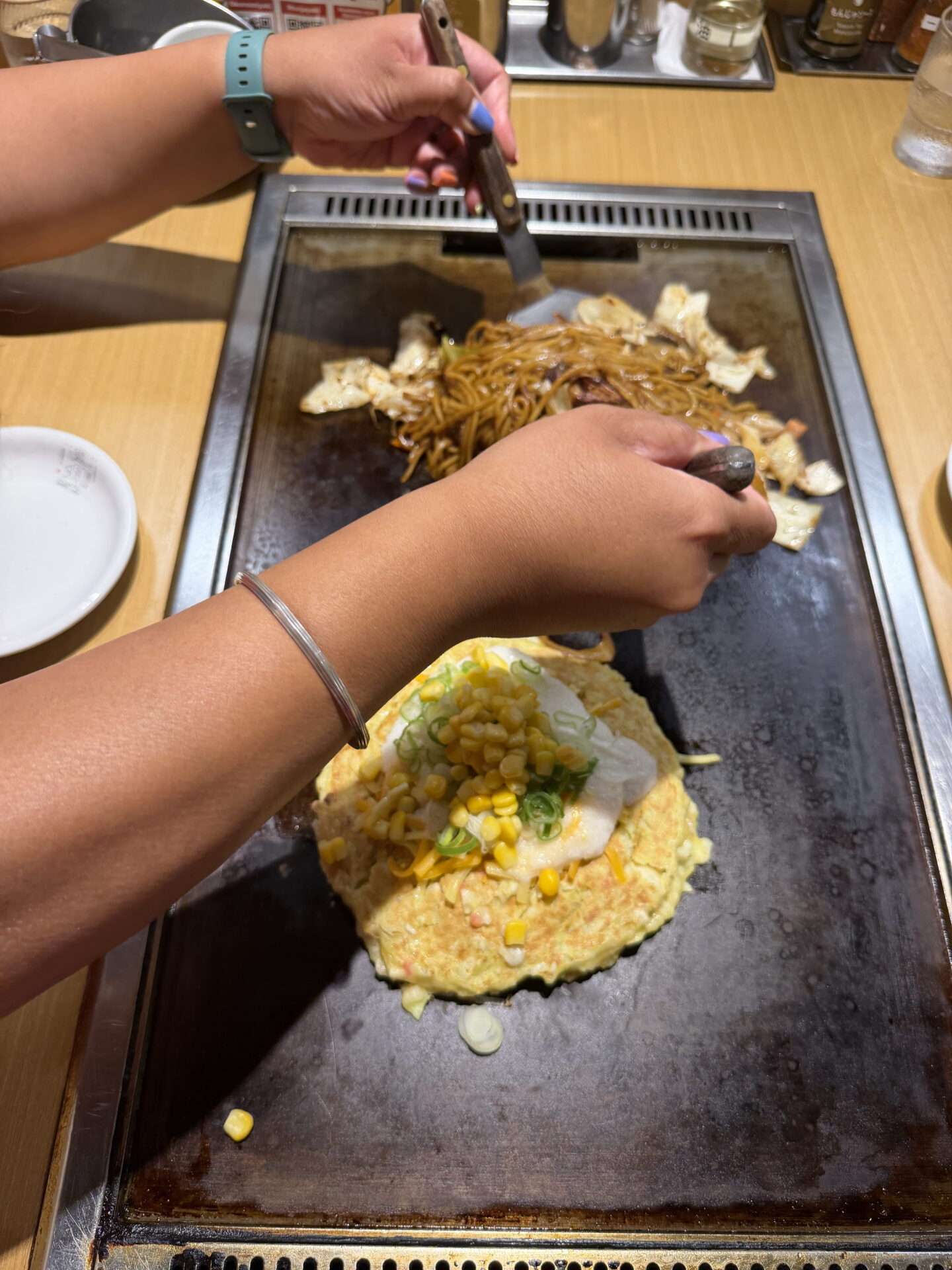 Close up of a woman using two metal spatulas to cook yakisoba noodles with cabbage on a Japanese teppan hotplate. Beside the noodles is a golden okonomiyaki pancake topped with sweetcorn, spring onions and a creamy white sauce.