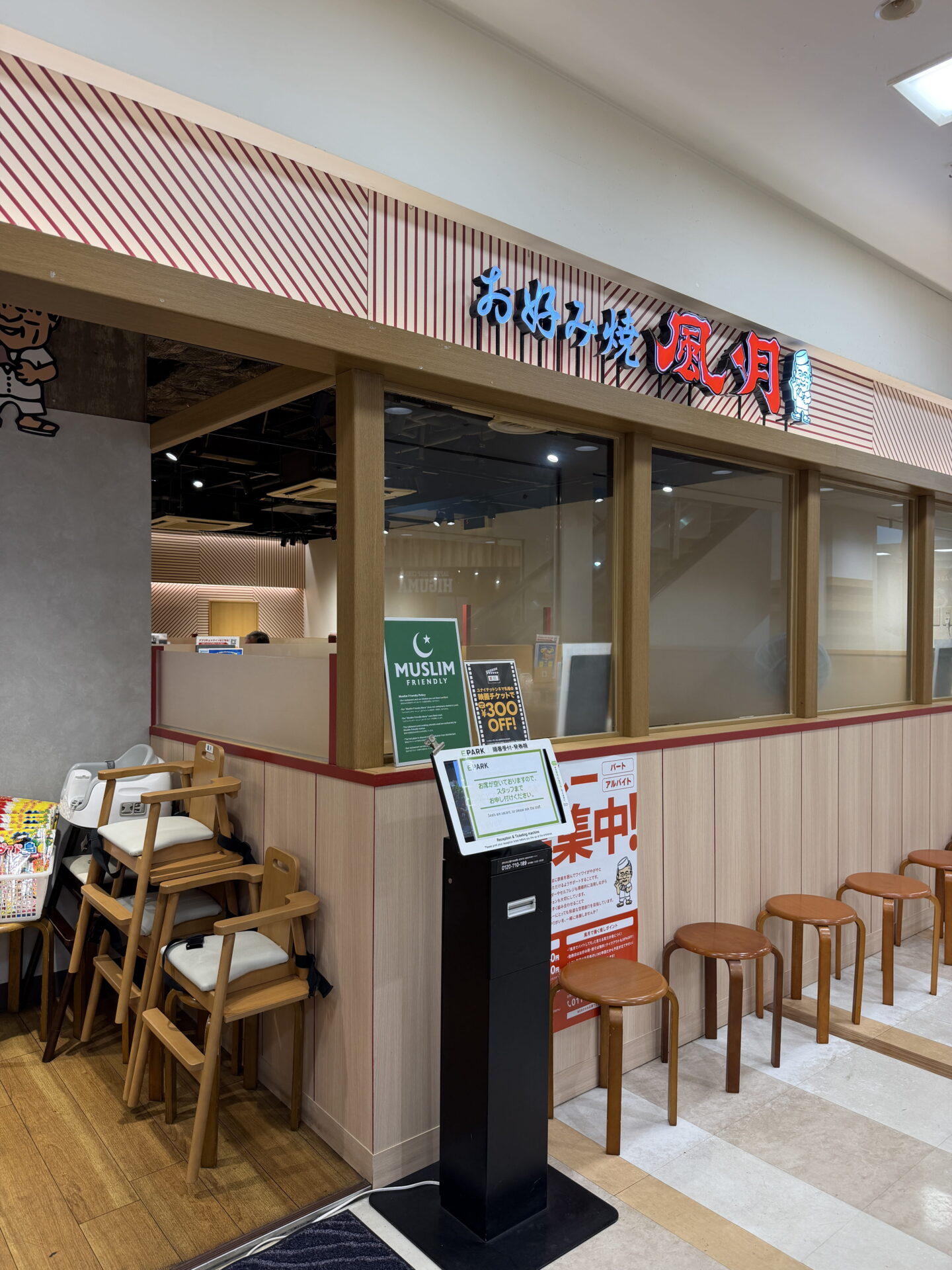 Entrance to a Japanese okonomiyaki restaurant with an illuminated sign reading お好み焼 風月 above large glass windows. A green sign reading "MUSLIM FRIENDLY" stands beside a self service check in kiosk, with wooden stools and stacked high chairs nearby.