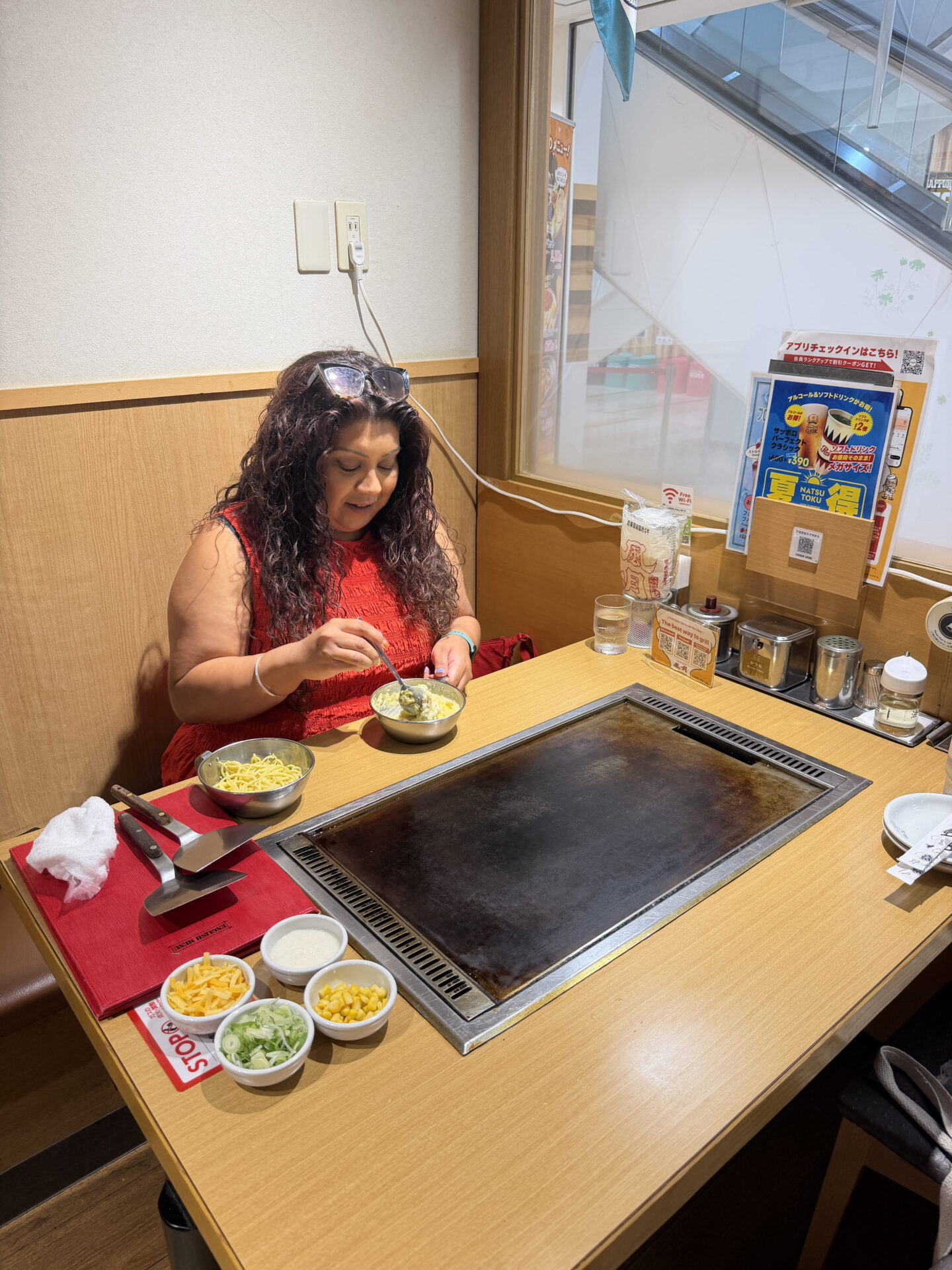 Nishi in a red sleeveless top mixes okonomiyaki batter in a metal bowl at a Japanese restaurant table with a built in hotplate. Small bowls of shredded cheese, sweetcorn, spring onions and sauce are arranged beside the grill ready for cooking.