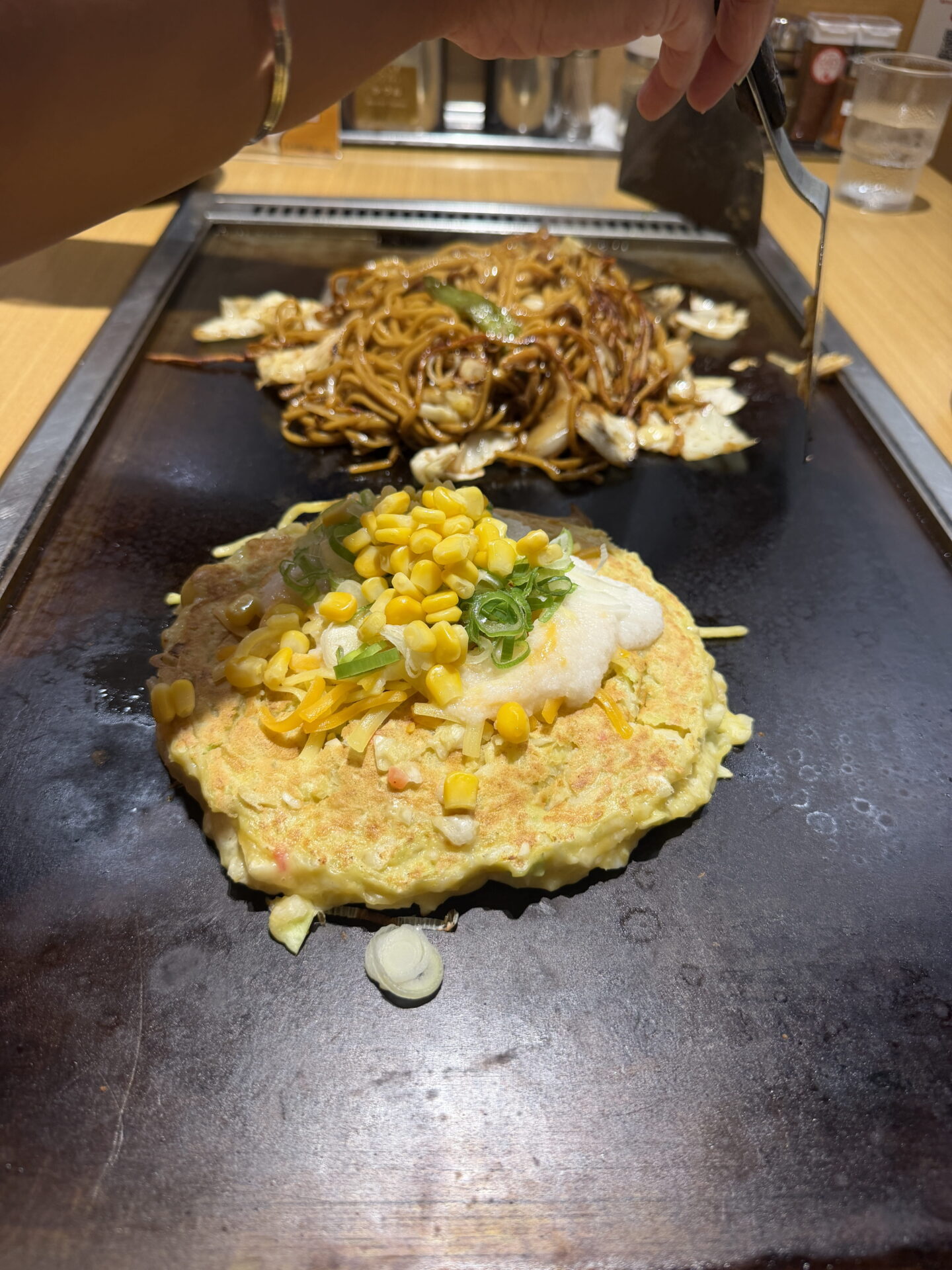 A golden Japanese okonomiyaki pancake topped with sweetcorn, spring onions, shredded cheese and creamy white sauce cooks on a tabletop hotplate. Behind it, yakisoba noodles mixed with cabbage and vegetables are being stirred with metal spatulas.