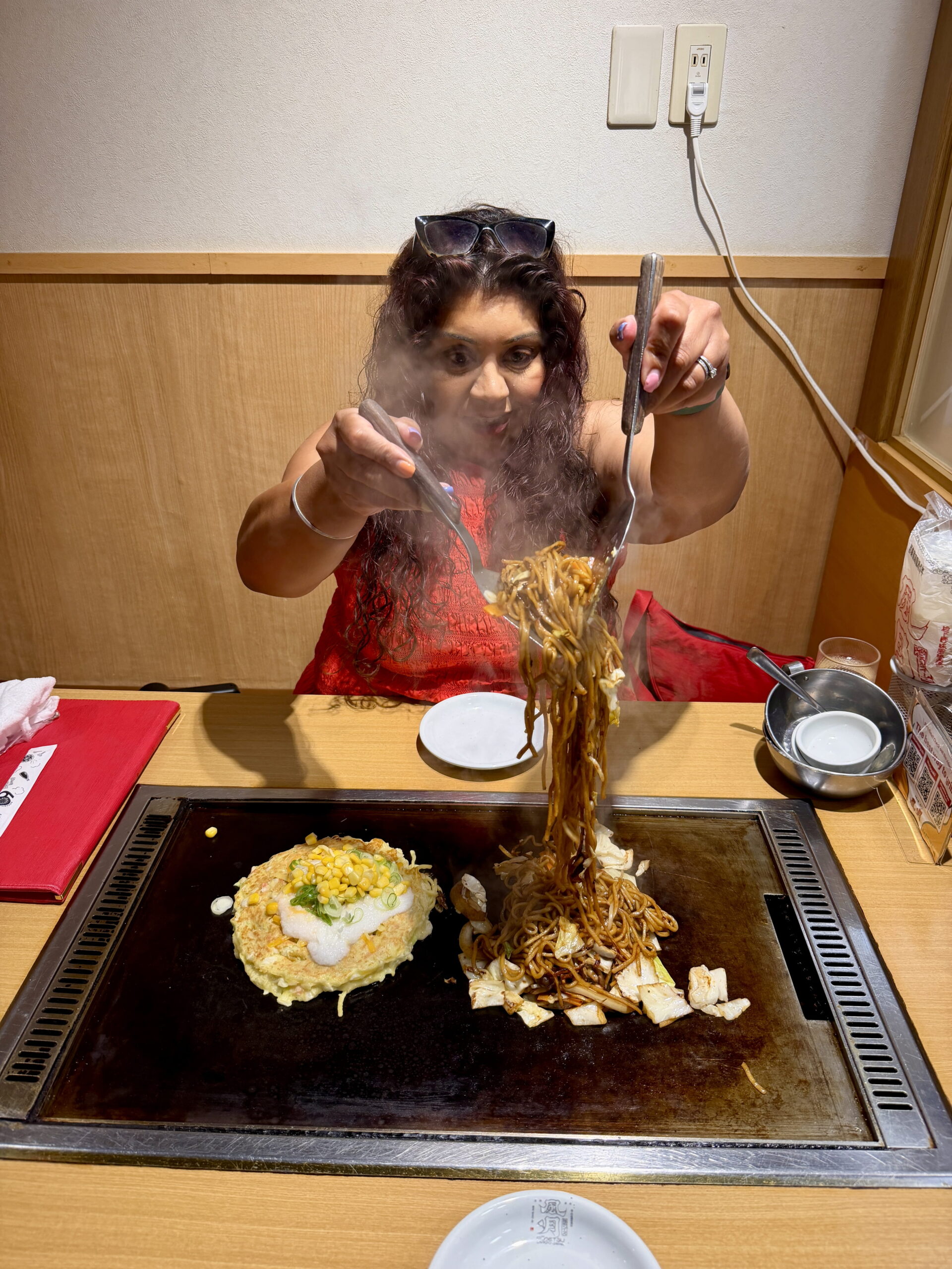 Nishi in a bright red top lifts a steaming portion of Japanese yakisoba noodles high above a tabletop hotplate using two metal spatulas. Beside the noodles sits a freshly cooked okonomiyaki pancake topped with sweetcorn, spring onions and white sauce.