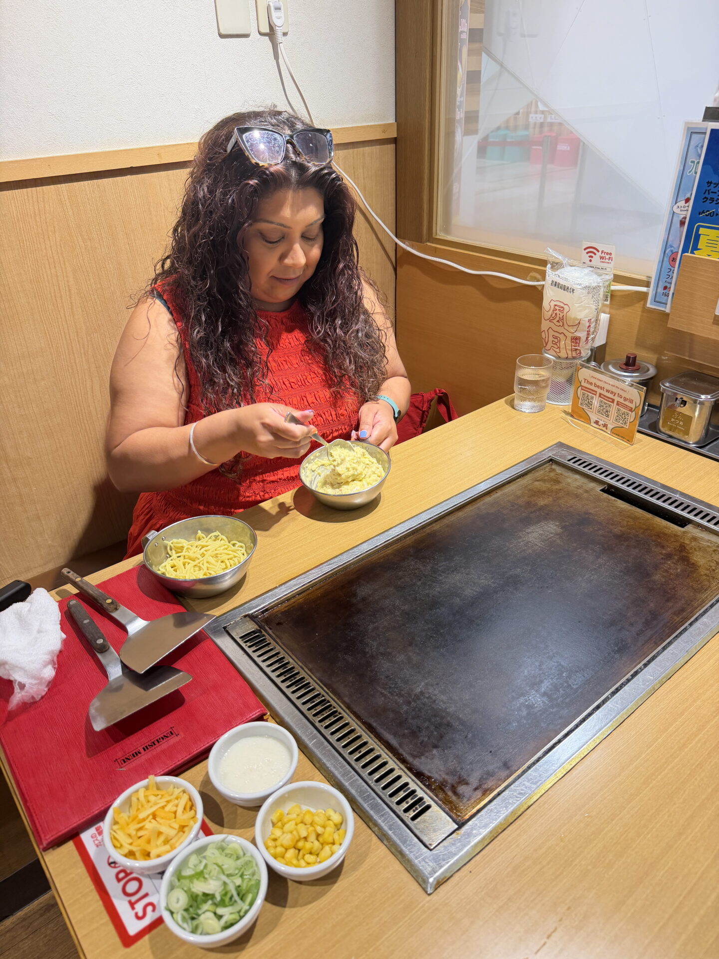 Nishi with long curly dark hair and sunglasses resting on her head mixes okonomiyaki batter in a metal bowl beside an empty tabletop hotplate. Bowls of sweetcorn, spring onions, shredded cheese and sauce are laid out on the wooden table