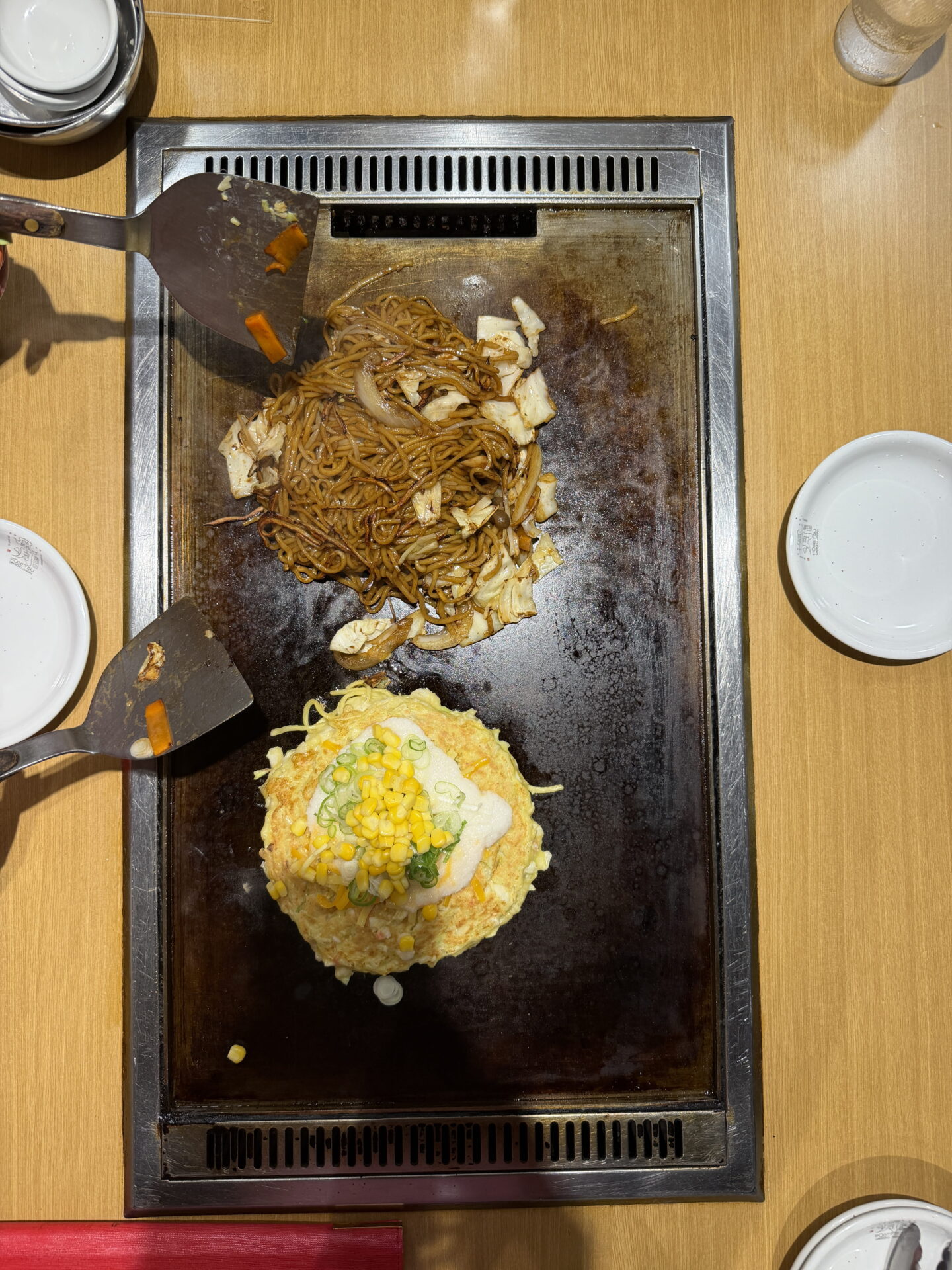 Overhead view of a Japanese tabletop hotplate with a round okonomiyaki pancake topped with sweetcorn, spring onions and white sauce beside a portion of yakisoba noodles with cabbage and onions. Two metal spatulas rest beside the freshly cooked dishes.