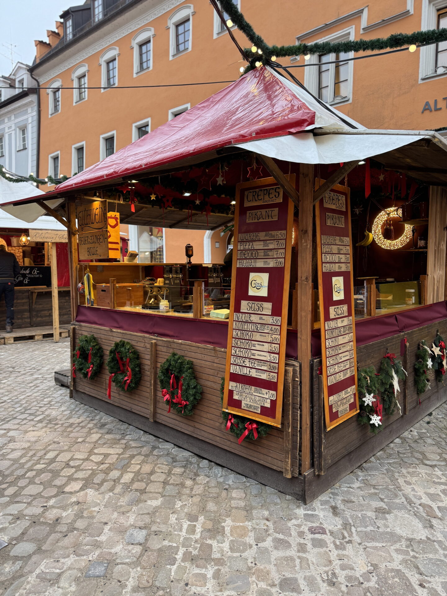 A wooden crêpe stall with a red and white canopy at a German Christmas market. Festive wreaths with red ribbons decorate the counter, while handwritten menus advertise sweet and savoury crêpes.