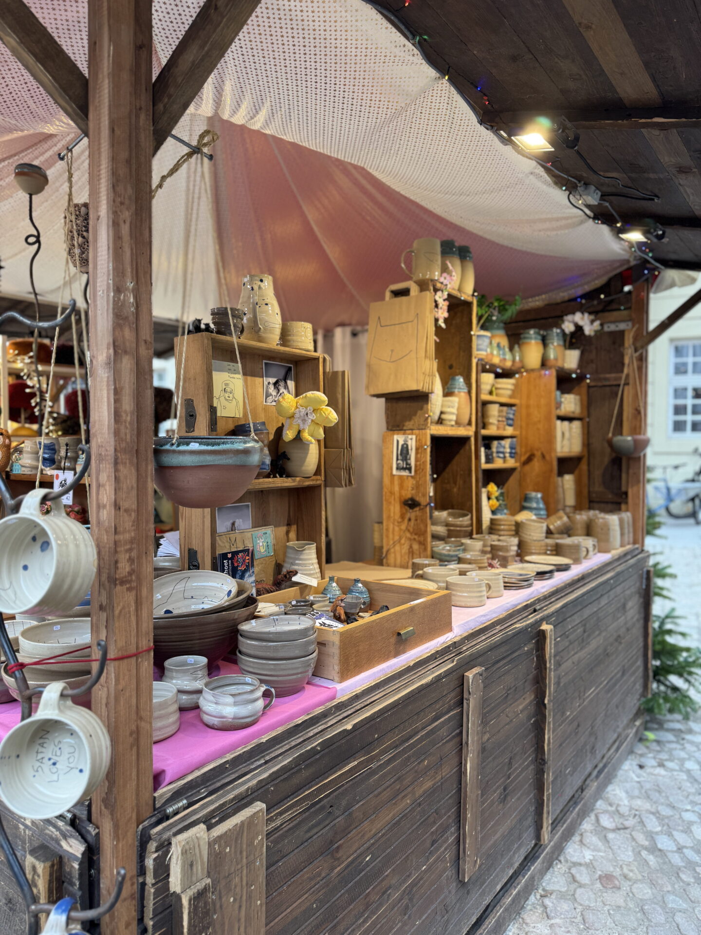 A wooden Christmas market stall displaying handmade ceramic bowls, mugs, plates and jugs in earthy colours. Pottery is arranged across wooden shelves and a pink tablecloth beneath a softly lit canopy.