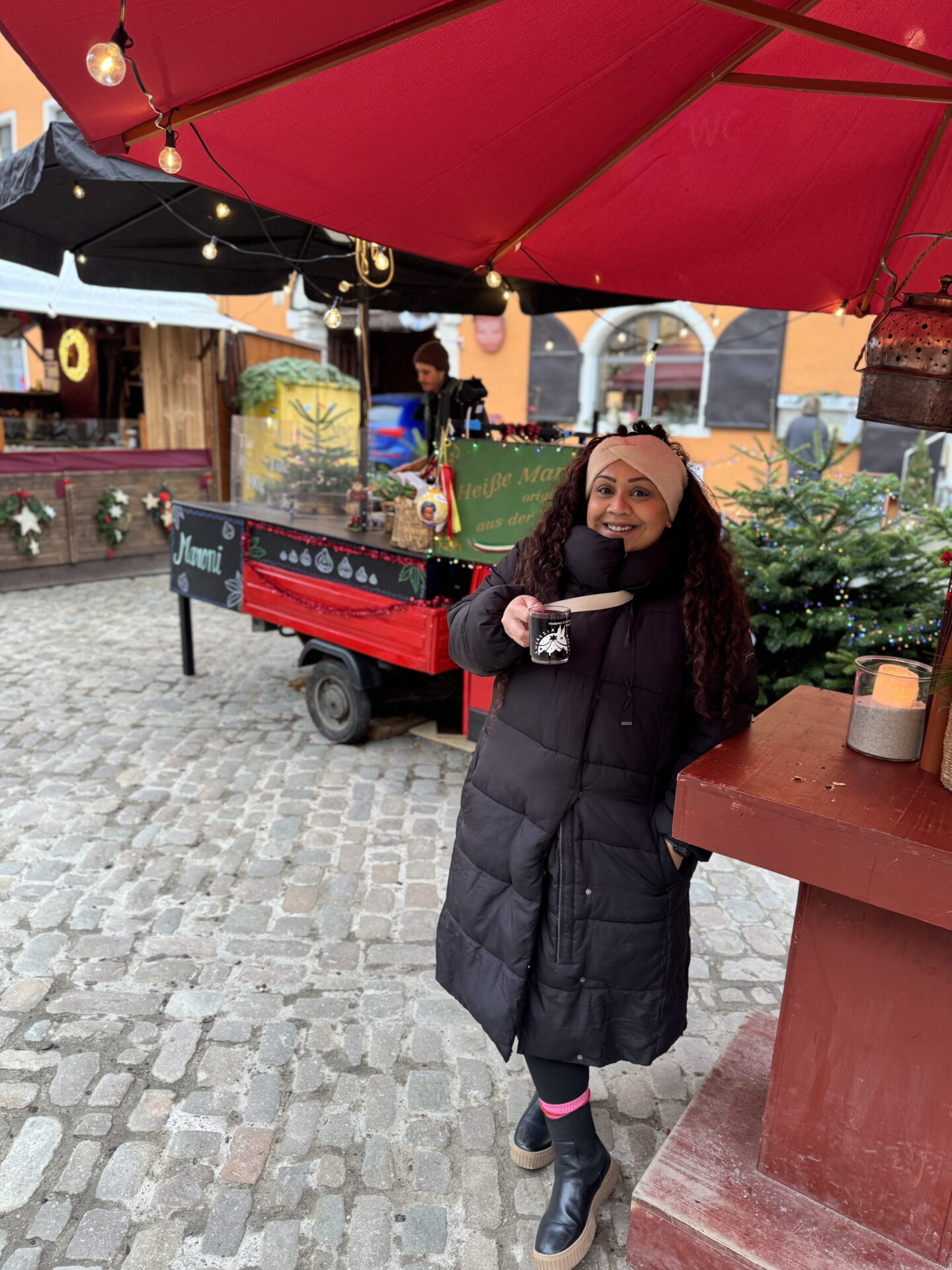 Nishi holds a small mug of hot drink beneath a large red umbrella. Behind her, a festive market stall and decorated Christmas trees brighten the cobbled square.