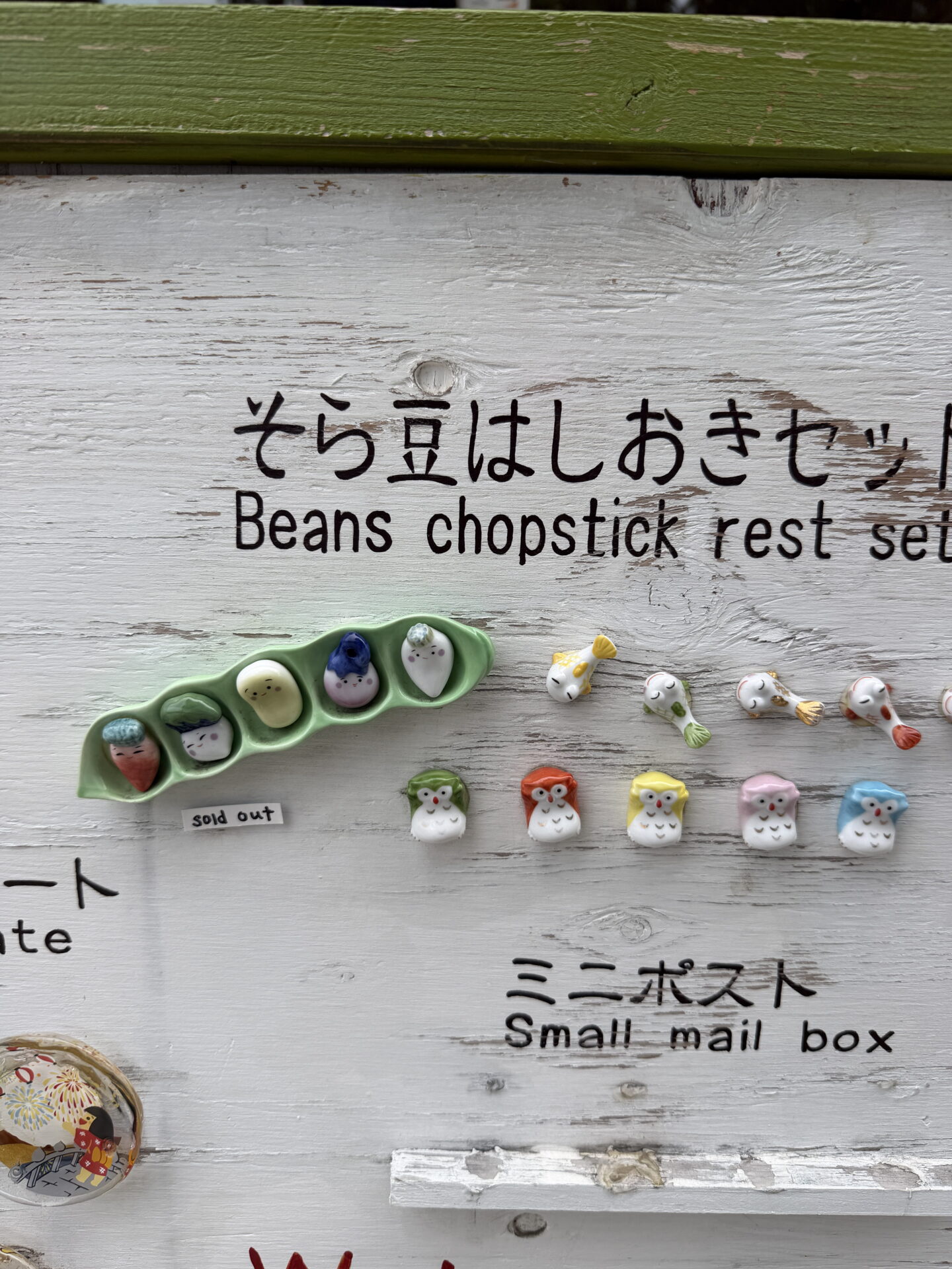 Colourful ceramic chopstick rests shaped like owls and small fish are displayed on a weathered white wooden board in a souvenir shop in Otaru. A green pea pod containing five miniature figures is labelled "sold out" while handwritten signs read "Beans chopstick rest set" and "Small mail box".