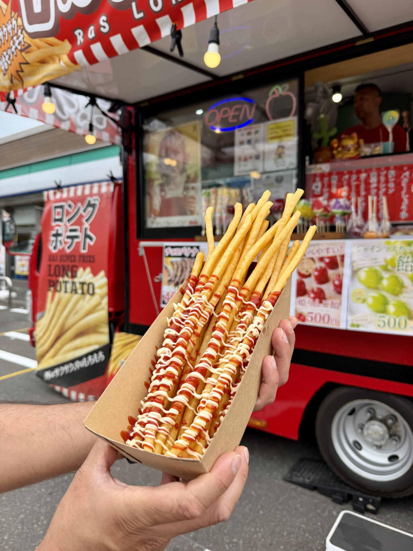 A person holds a cardboard tray filled with extra long golden potato fries drizzled with red ketchup and white mayonnaise. Behind the fries is a bright red Japanese street food van with a sign advertising "LONG POTATO" and a glowing "OPEN" sign.