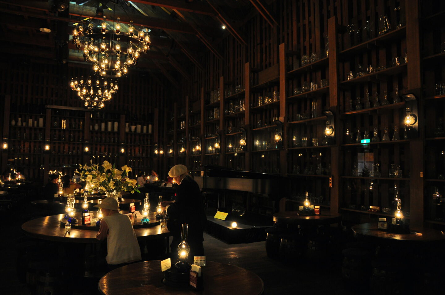 A dimly lit café in Otaru with towering wooden shelves displaying hundreds of glass ornaments and vessels. Glowing oil lamps illuminate the round wooden tables where visitors sit beneath elaborate chandeliers made from glass bottles.