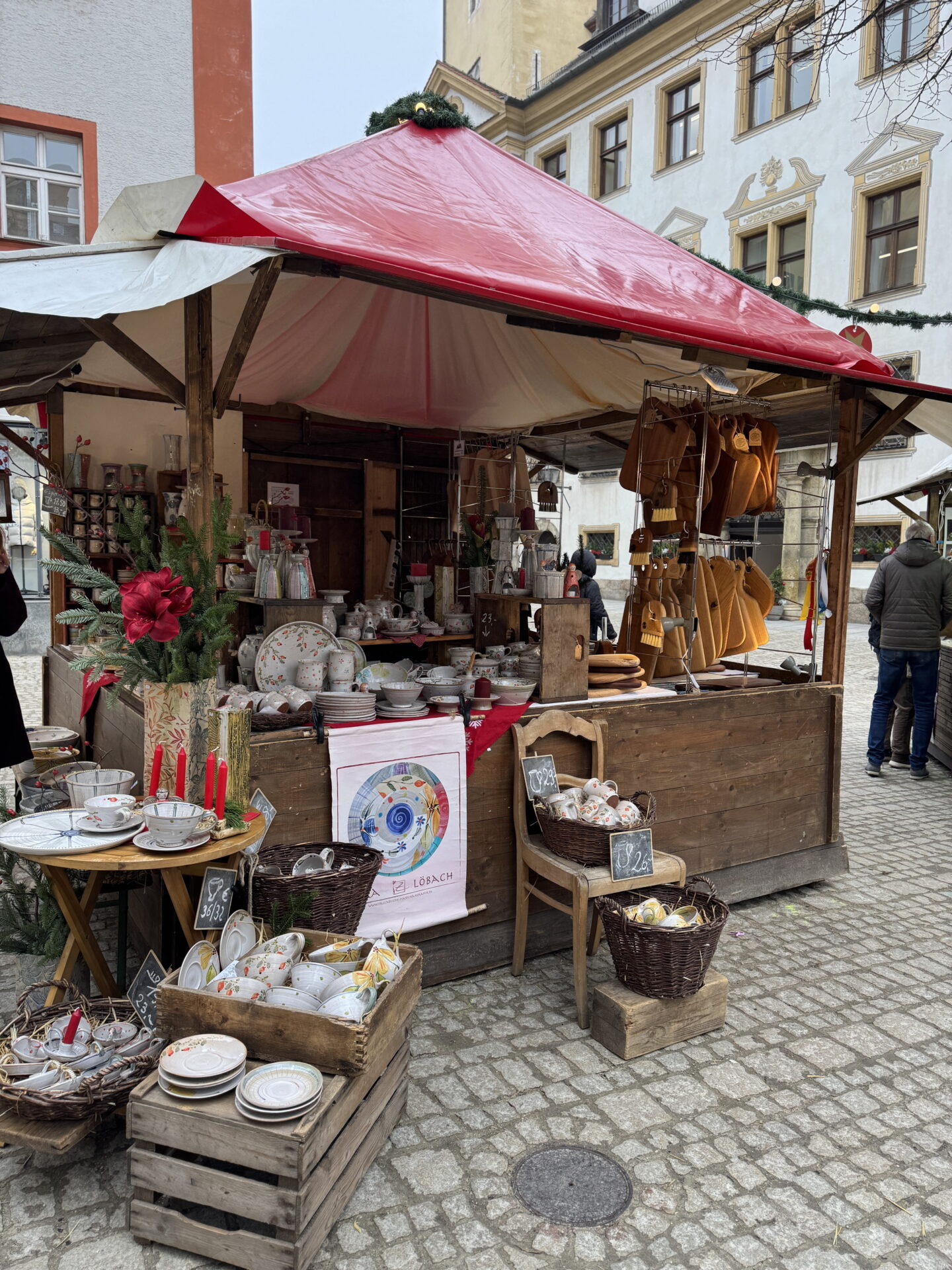 A wooden Christmas market stall with a red canopy displays colourful ceramic plates, cups, bowls and handmade gifts. Baskets of pottery and hanging mustard coloured mittens surround the stall, decorated with evergreen branches and red candles.