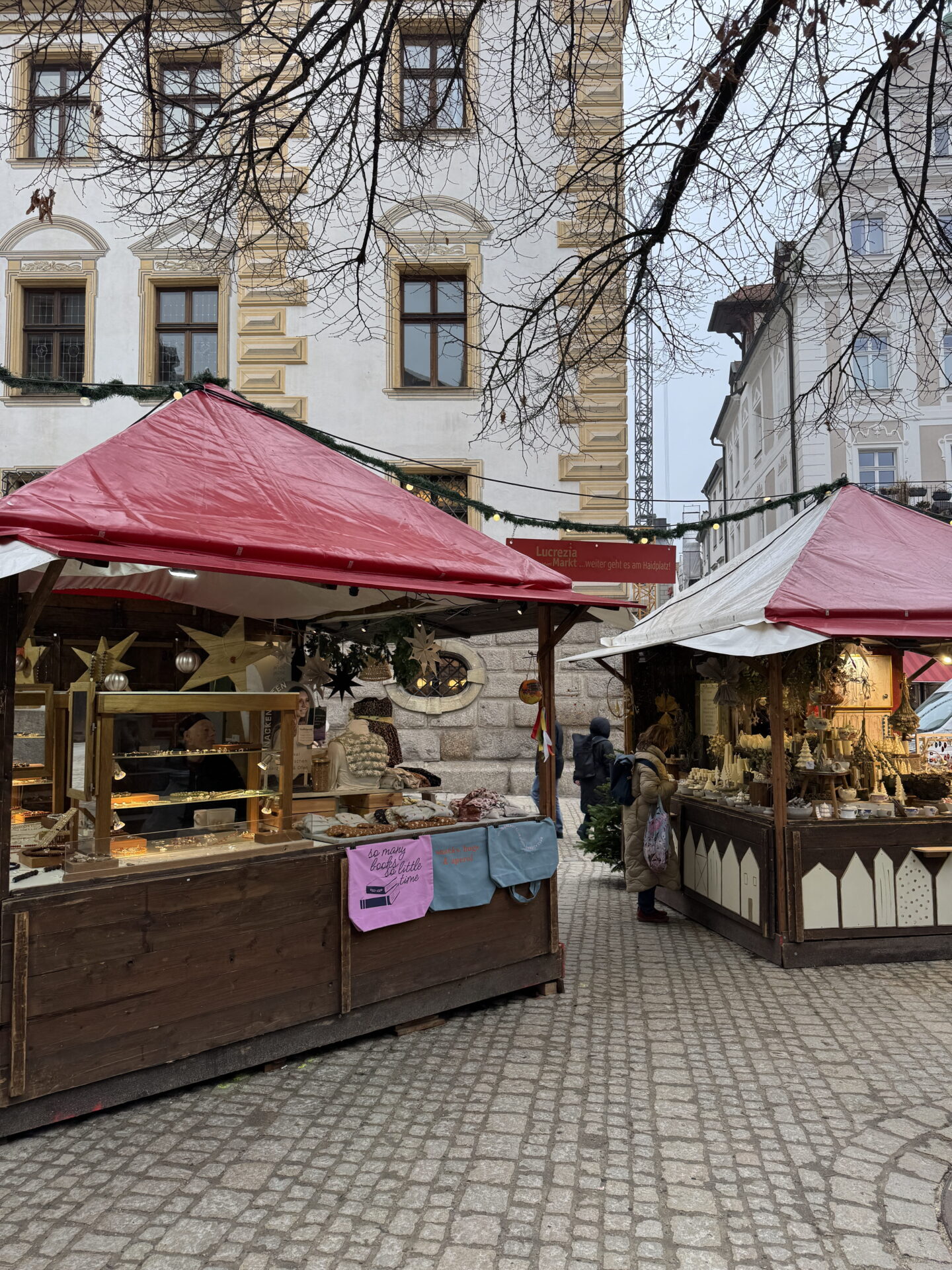 Two wooden Christmas market stalls with red and white roofs stand on a cobbled square surrounded by historic buildings. One stall displays baked goods while the other sells festive decorations, with shoppers browsing beneath strings of lights.