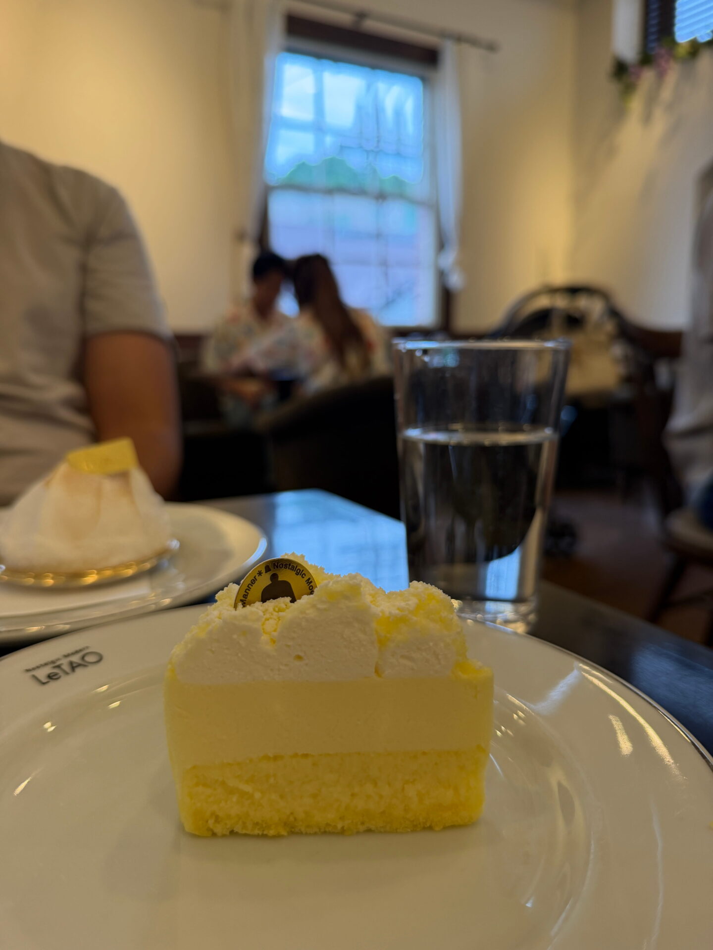 A slice of layered cheesecake topped with fluffy whipped cream sits on a white LeTAO plate at a café in Otaru. A glass of water and another plated dessert sit on the table with customers and a curtained window in the background.