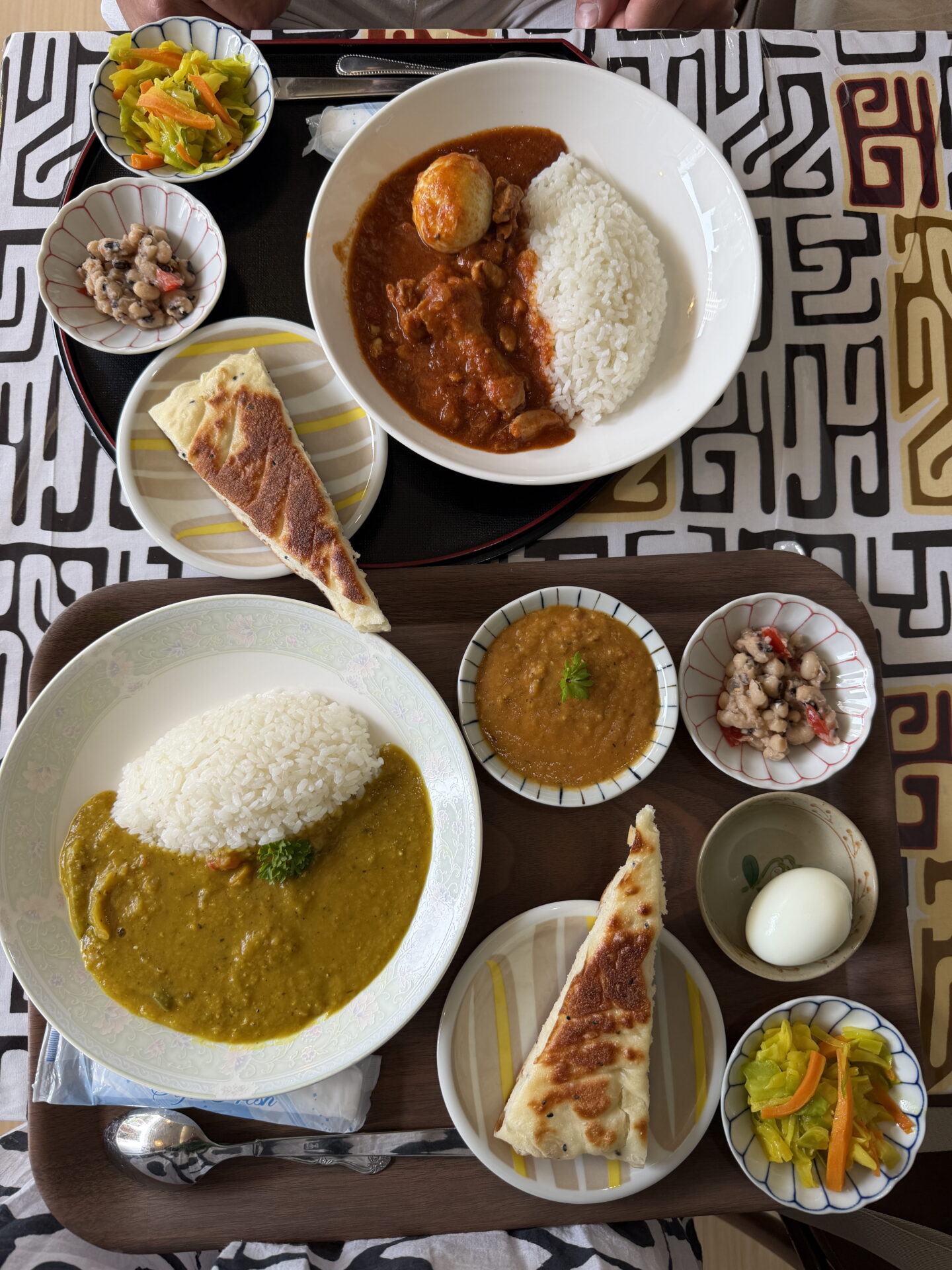 Overhead view of two Japanese curry meals served on separate trays, one with a reddish brown curry containing a boiled egg and the other with a yellow curry, both accompanied by white rice. Side dishes include pieces of naan bread, lentil curry, bean salad, pickled vegetables and a boiled egg.