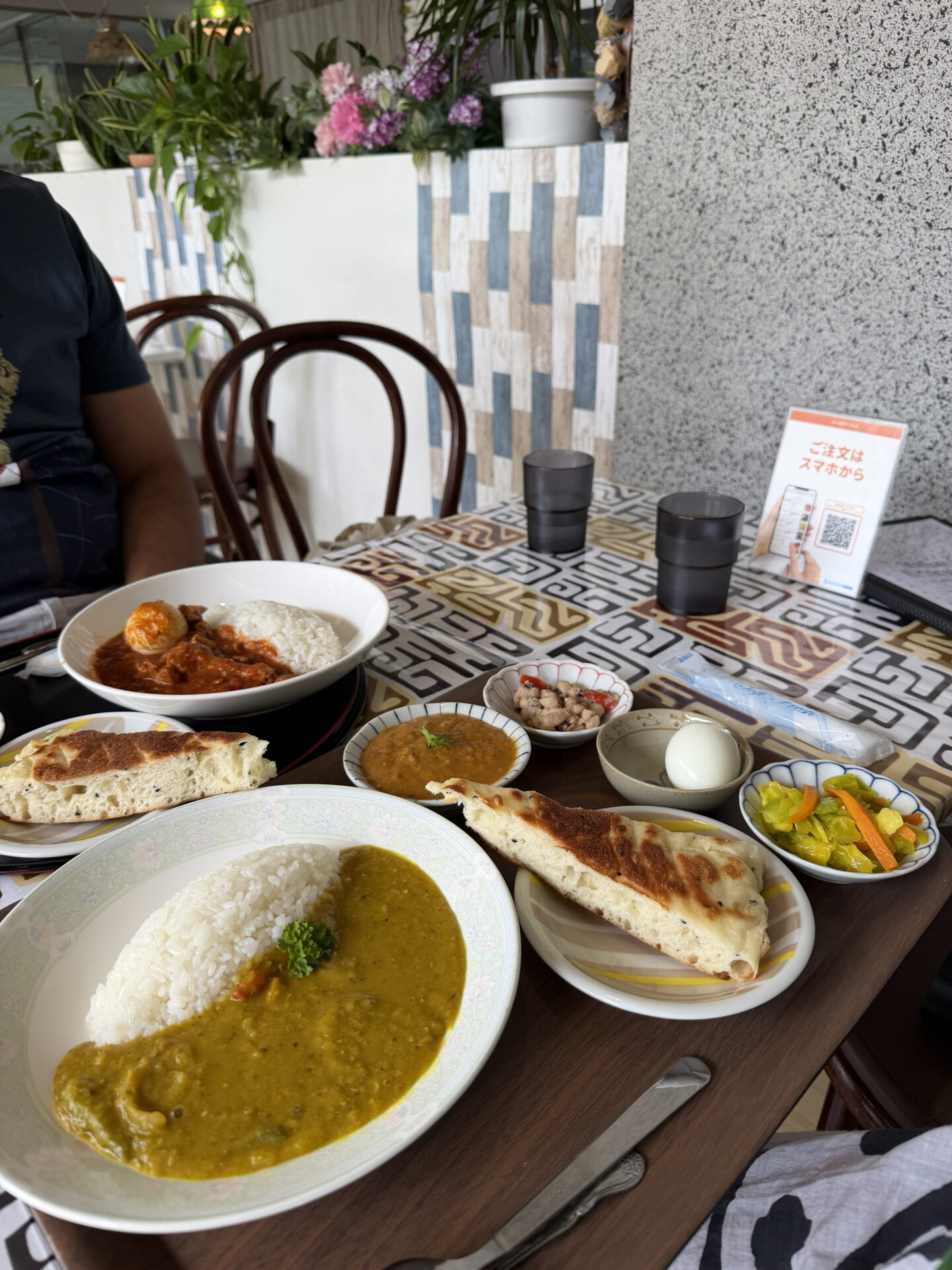 Two Japanese curry meals arranged on a restaurant table with patterned tiles, featuring bowls of red and yellow curry served with white rice and naan bread. Small side dishes of lentils, beans, pickled vegetables and a boiled egg complete the spread.