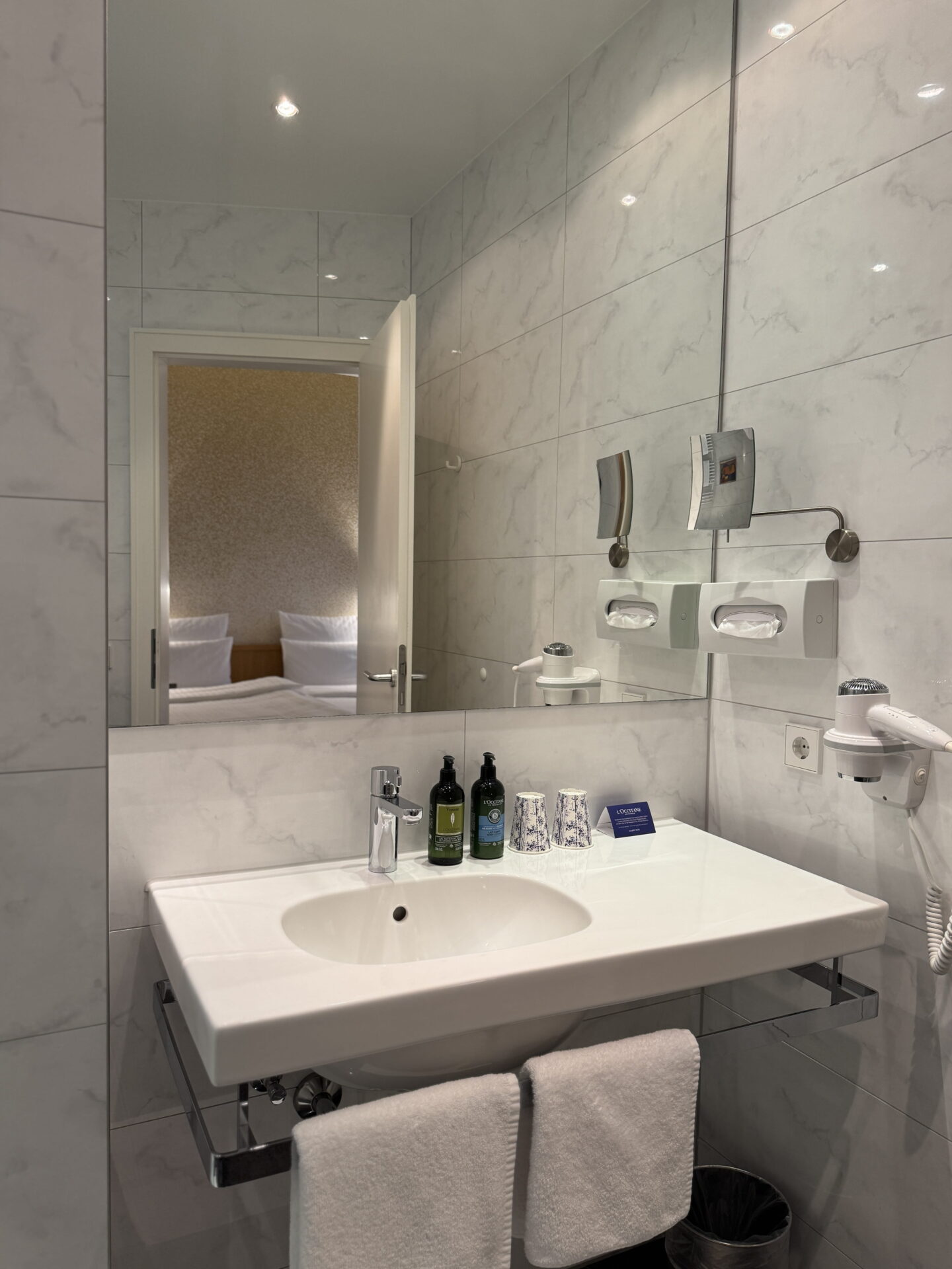 A modern hotel bathroom with pale marble effect tiles, a white rectangular sink and a large wall mirror reflecting the adjoining bedroom. Green L Occitane toiletries, patterned cups and a wall mounted hairdryer sit above neatly folded white hand towels.