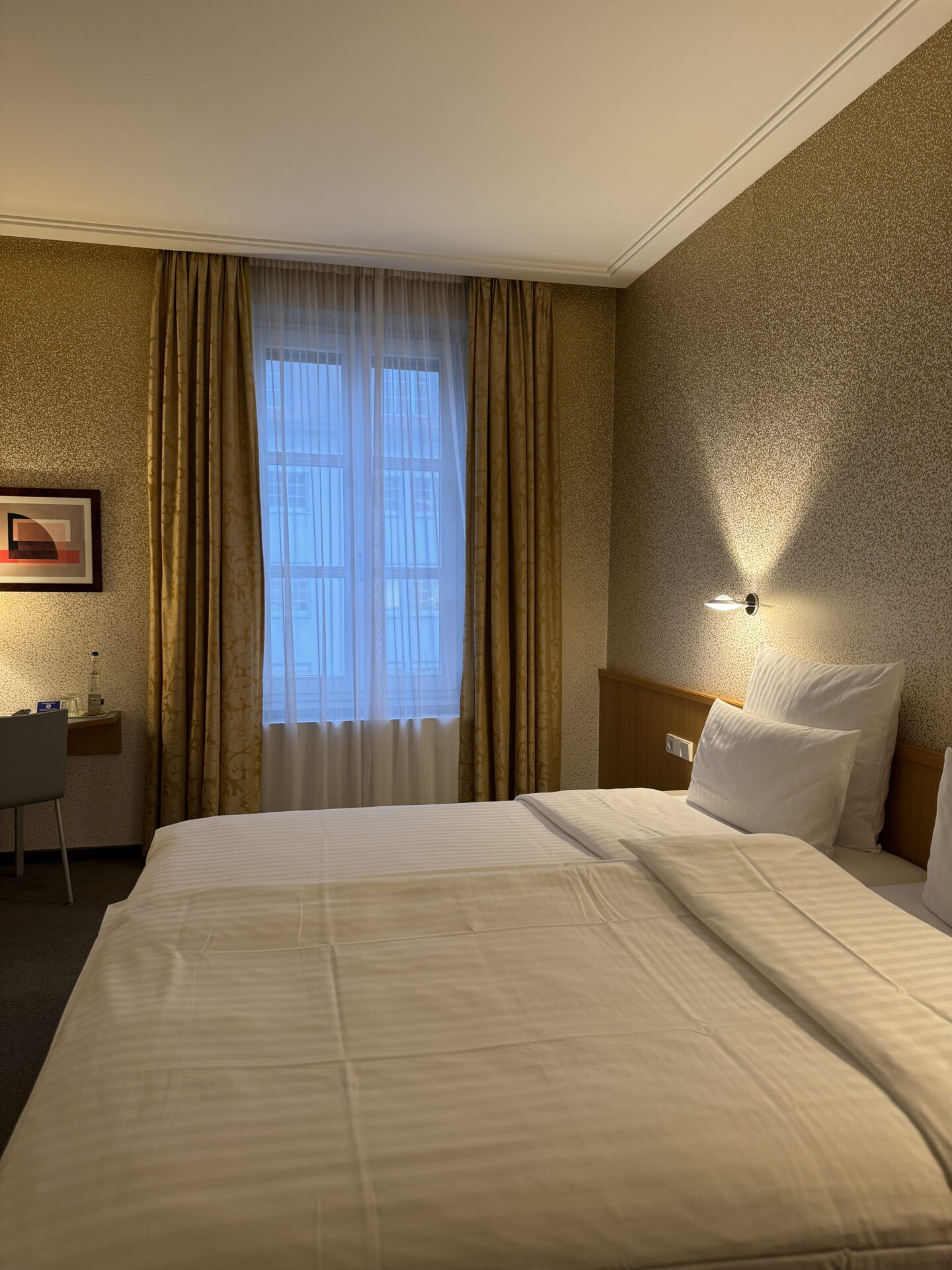 A hotel bedroom with a neatly made bed dressed in white striped bedding and plump pillows against a wooden headboard. Gold patterned curtains frame a tall window, with a small desk and chair tucked into the corner.