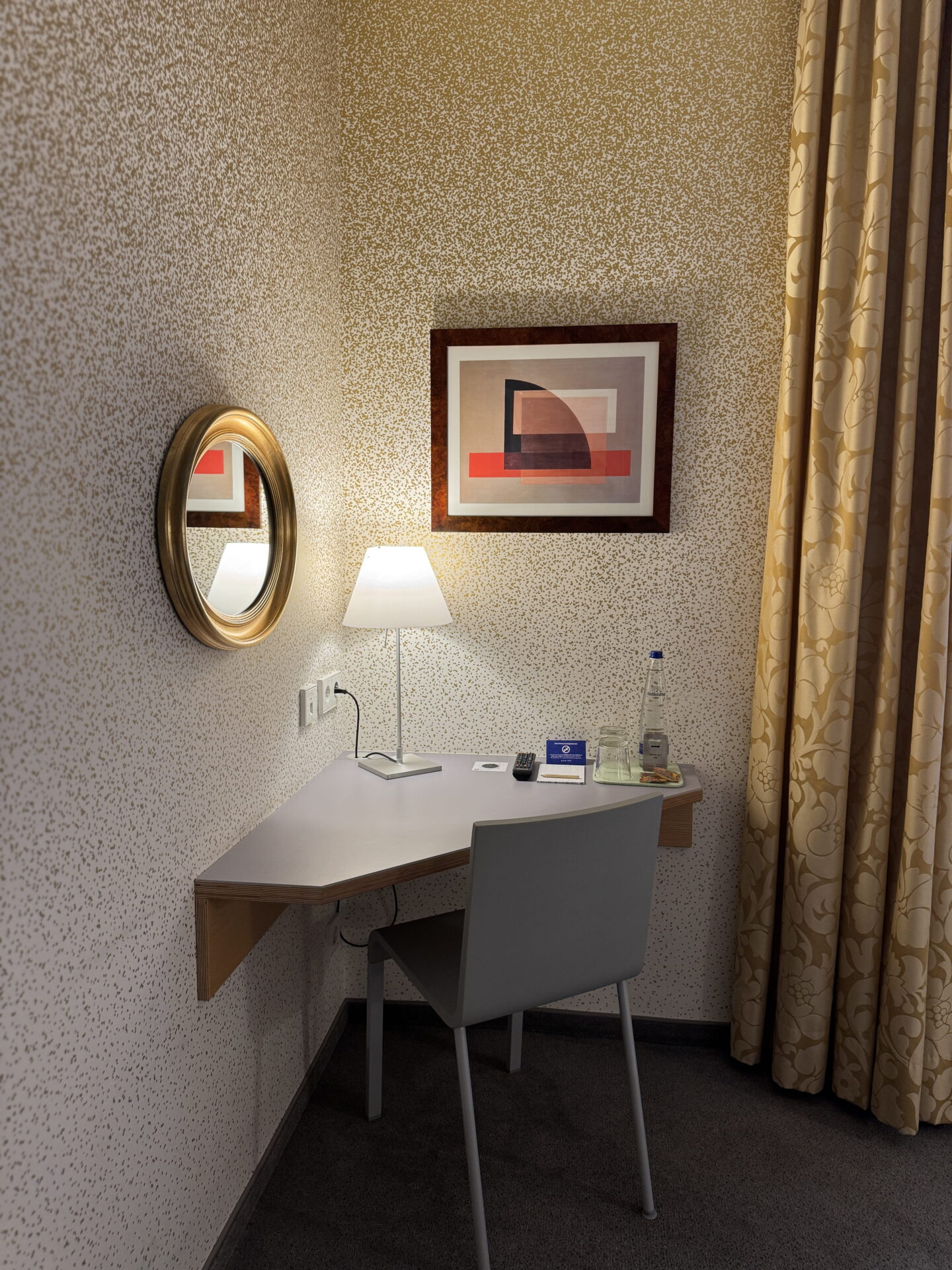 A compact hotel room workspace with a grey corner desk, matching chair and white table lamp beneath a framed abstract print. A round gold framed mirror hangs on the patterned wall, with bottled water and glasses arranged on the desk.