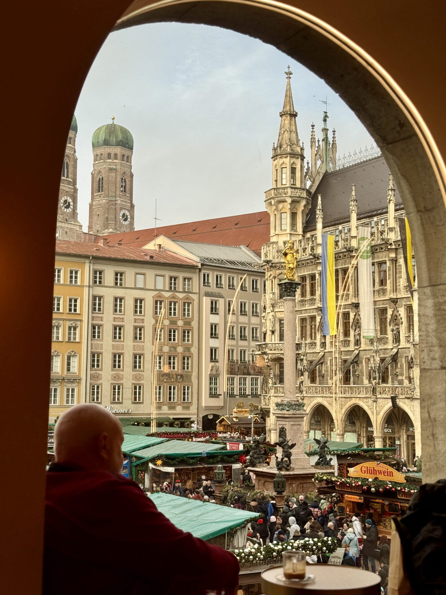 View of Munich Christmas Market in Marienplatz through a large stone archway, with crowds browsing green roofed stalls around the golden Marian column. The twin green domes of Frauenkirche and the Gothic New Town Hall rise above the square.