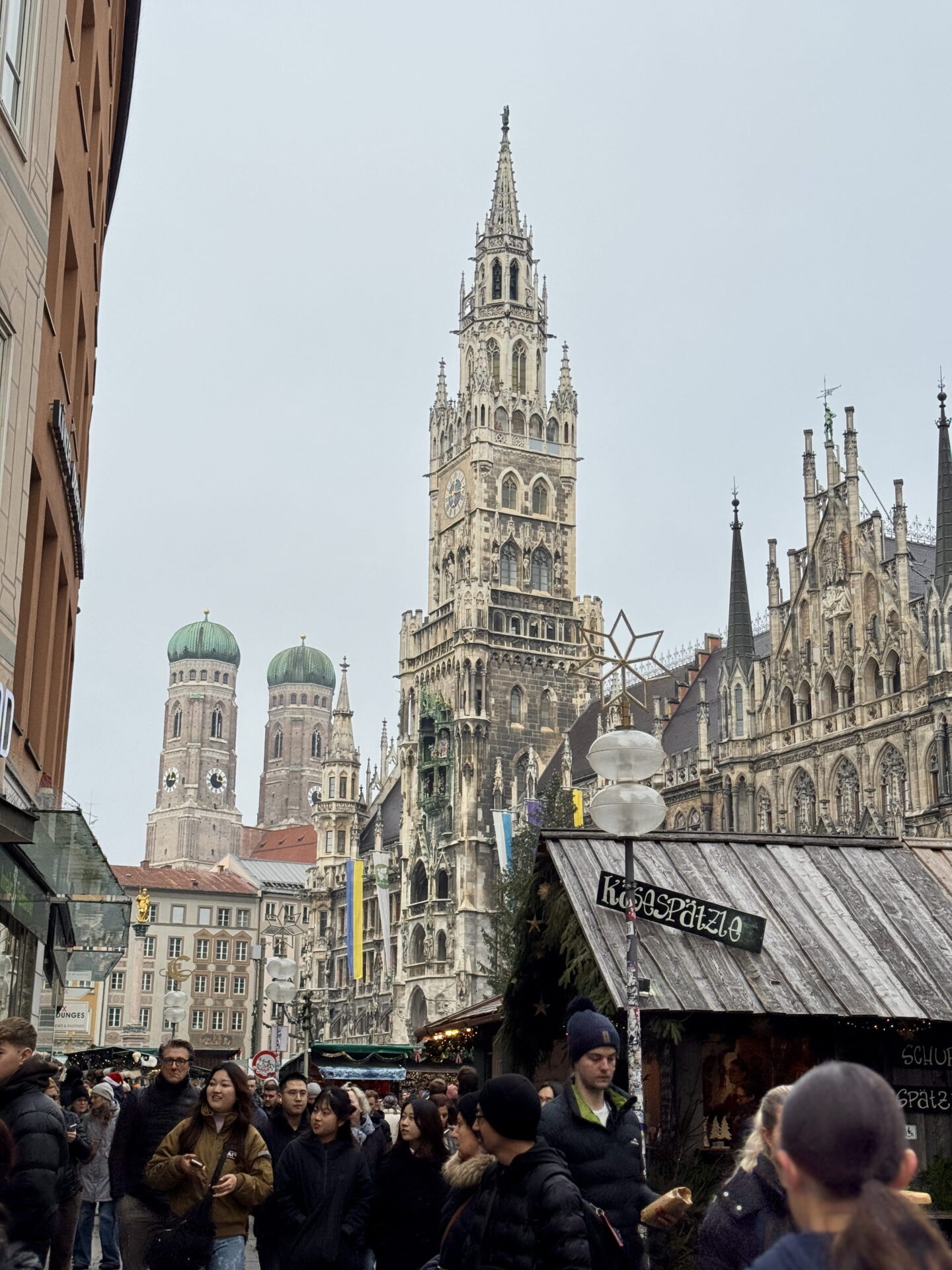 Crowds of visitors walking through Munich Christmas Market in Marienplatz, with wooden food stalls lining the street. The Gothic clock tower of the New Town Hall and the twin green domes of Frauenkirche dominate the skyline.
