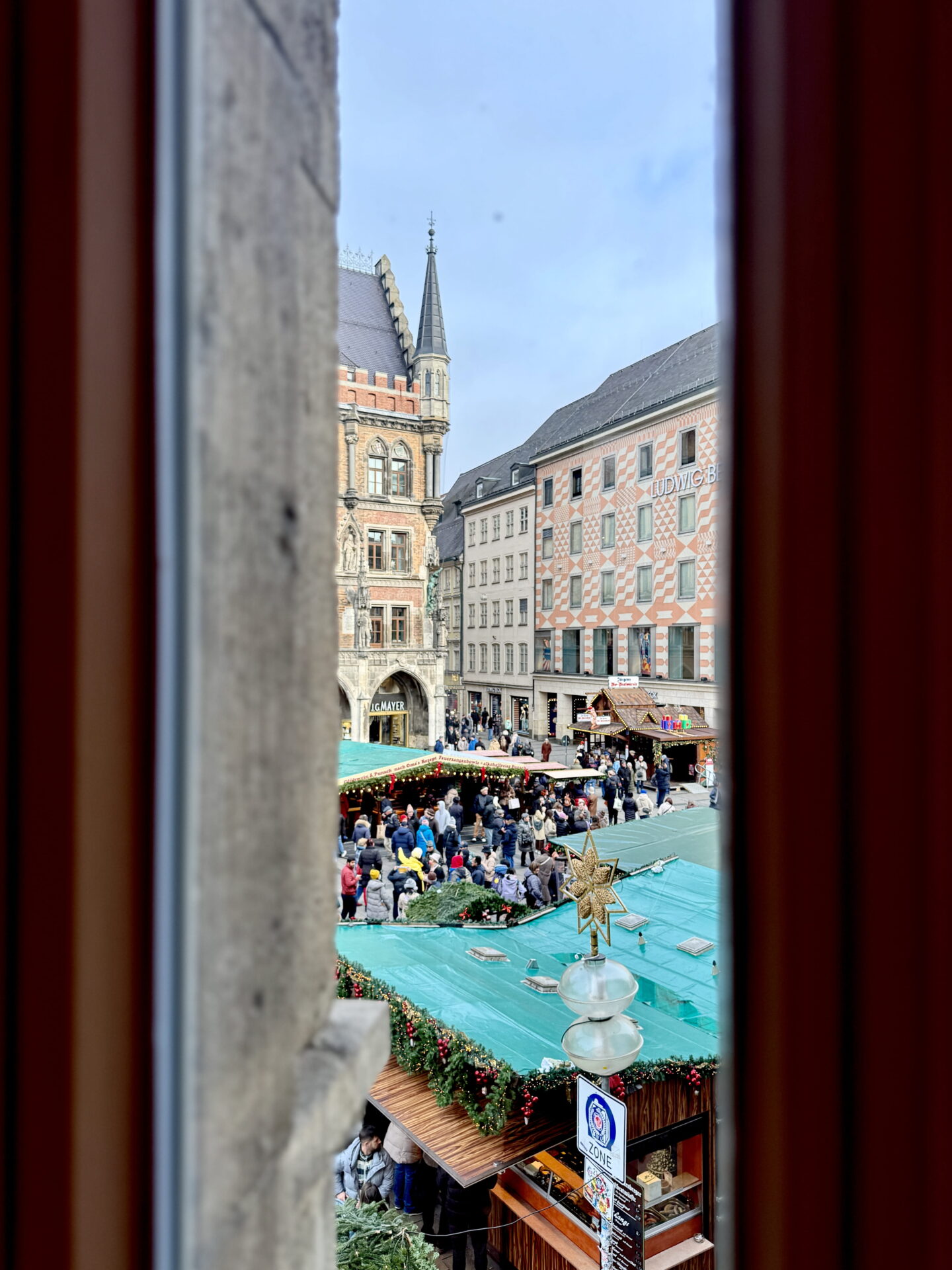 Elevated view of Munich Christmas Market through a narrow stone window, overlooking crowds gathered around wooden stalls with green roofs and festive garlands. Historic buildings surround the busy square, including a pink and white patterned building.