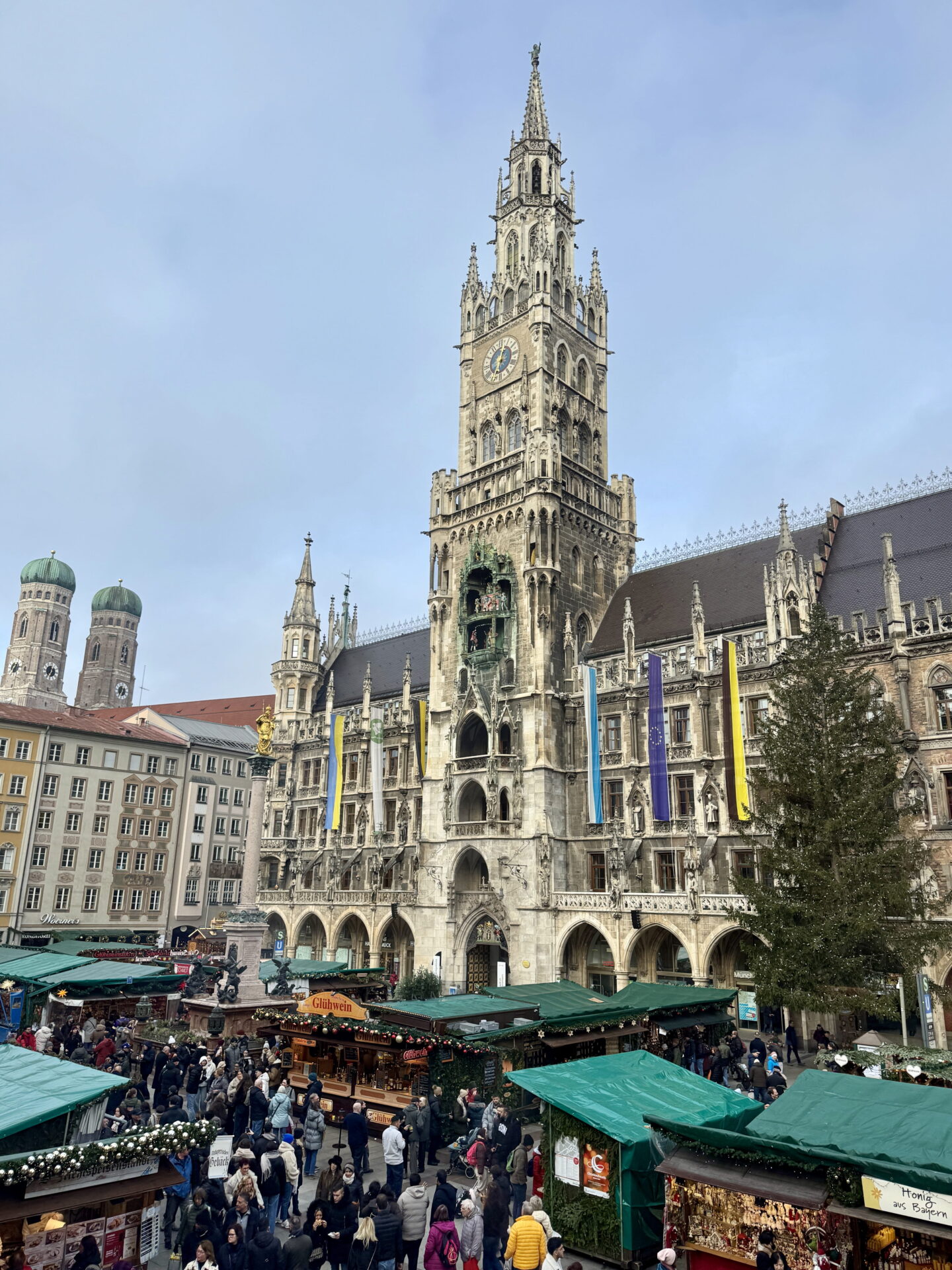 The Gothic New Town Hall in Munich towers above the busy Marienplatz Christmas Market, with its ornate clock tower and colourful hanging banners. Visitors browse wooden stalls with green roofs beside a tall Christmas tree.