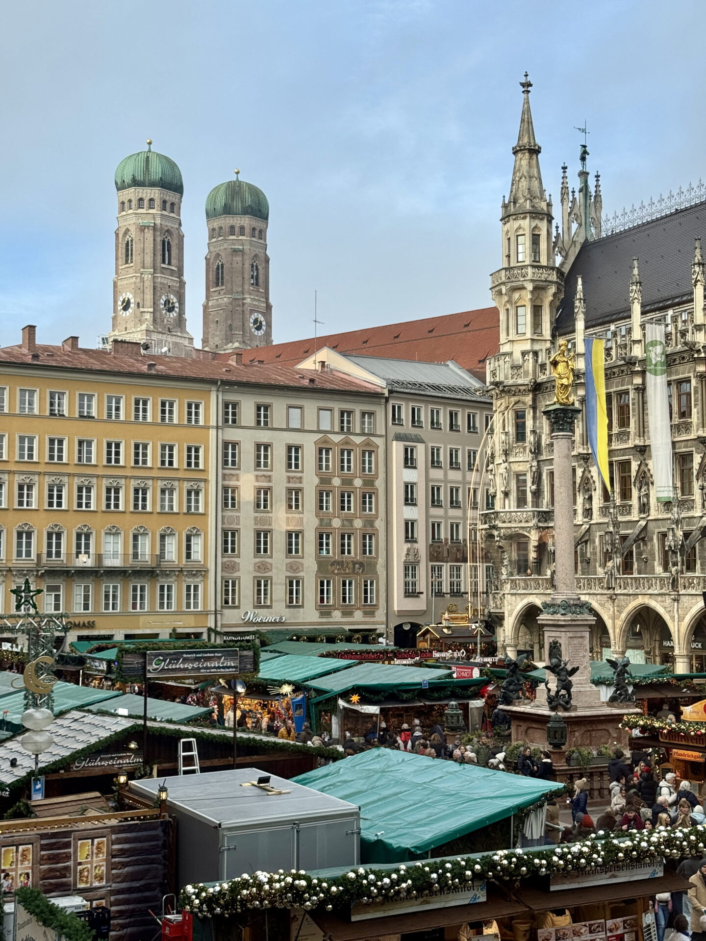 Elevated view across Munich Christmas Market in Marienplatz, with crowds browsing festive wooden stalls around the golden Marian column. The twin green domes of Frauenkirche rise above pastel coloured buildings beside the ornate New Town Hall.