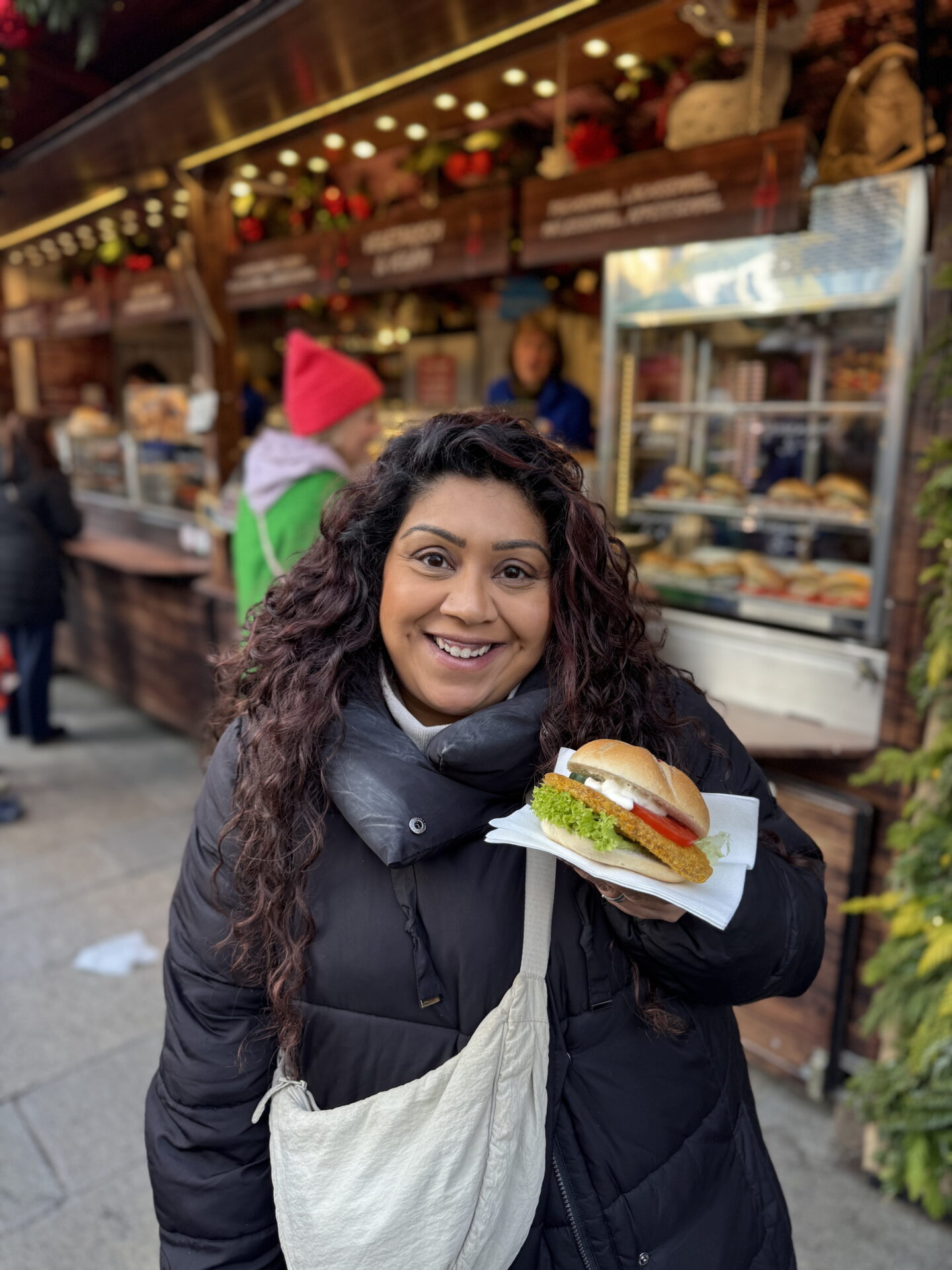 Nishi smiles at the camera while holding a bread roll filled with a golden crumbed fish fillet, lettuce, tomato and white sauce. She wears a black puffer coat and cream crossbody bag in front of a wooden Christmas market food stall displaying sandwiches.
