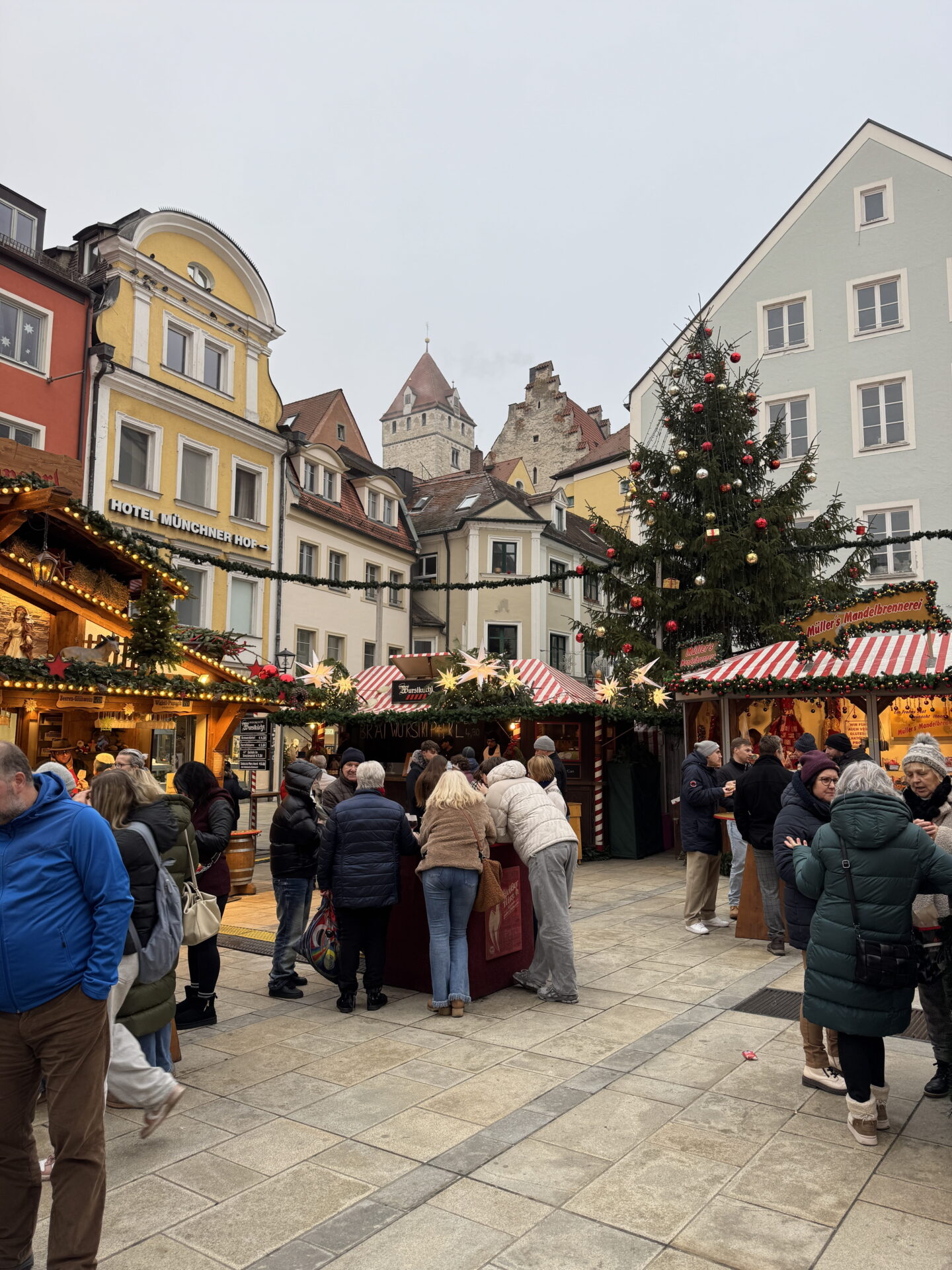 A tall Christmas tree decorated with red and gold baubles stands between illuminated market stalls in a busy German town square. Visitors in winter coats gather around food stalls, with colourful historic buildings and a stone tower in the background.