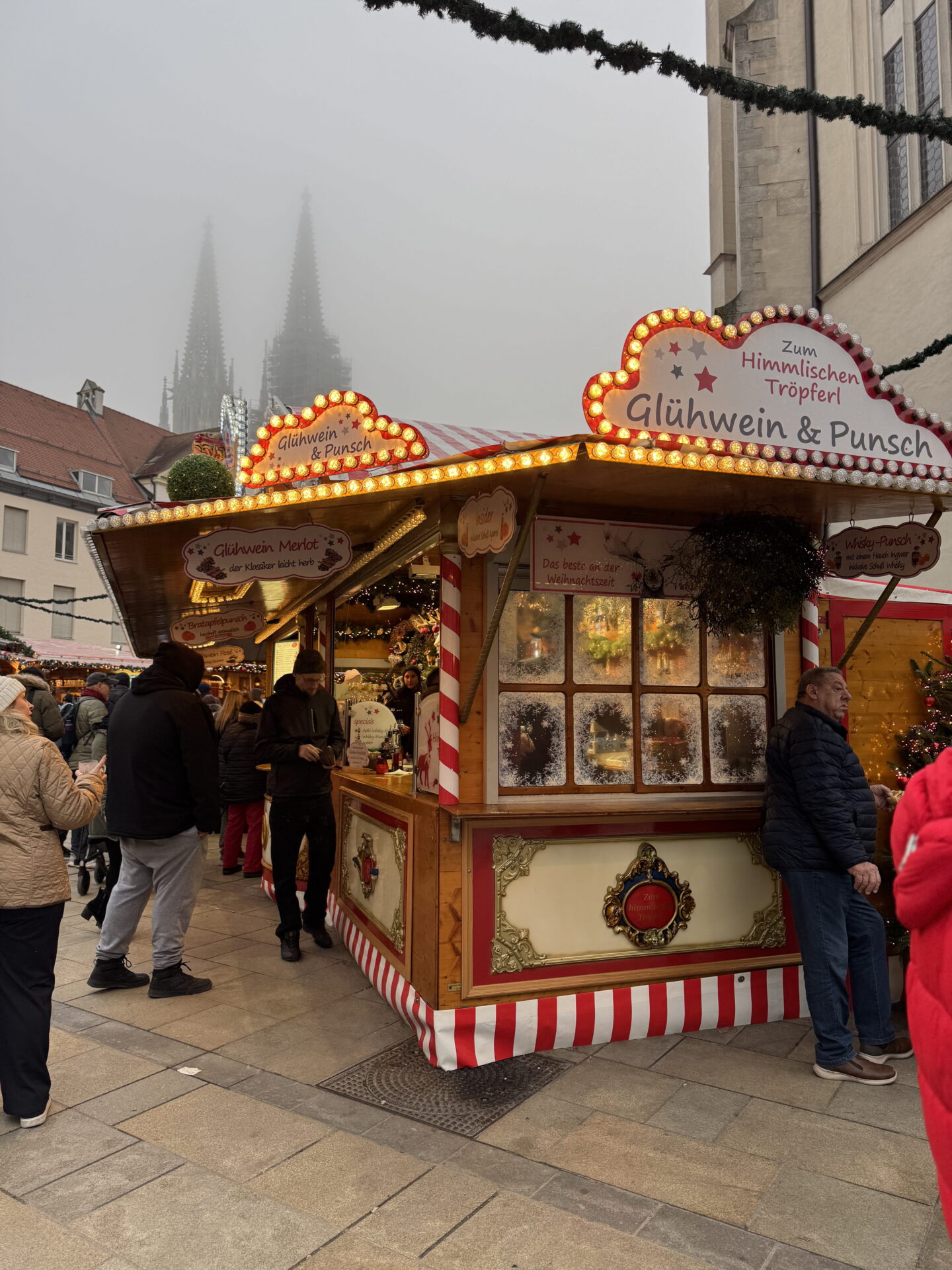 A brightly illuminated wooden drinks stall at a German Christmas market advertises Glühwein and Punsch beneath glowing signs. Visitors gather around the stall while the twin Gothic spires of a cathedral rise through the mist in the background.