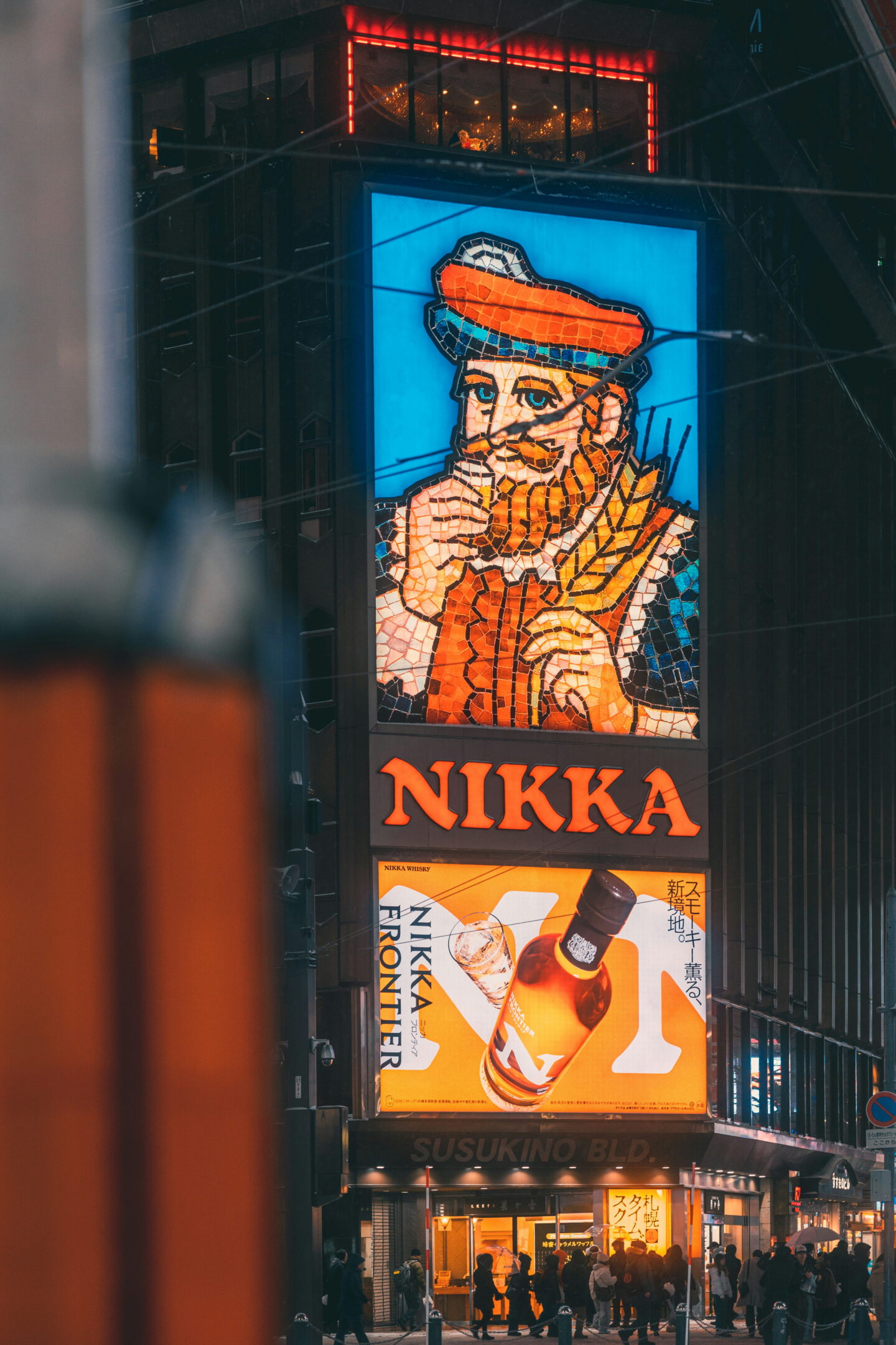 The famous illuminated Nikka whisky billboard in the Susukino entertainment district of Sapporo at night. A colourful mosaic portrait of a bearded man wearing a red hat sits above the glowing Nikka sign and an advertisement for Nikka Frontier whisky.