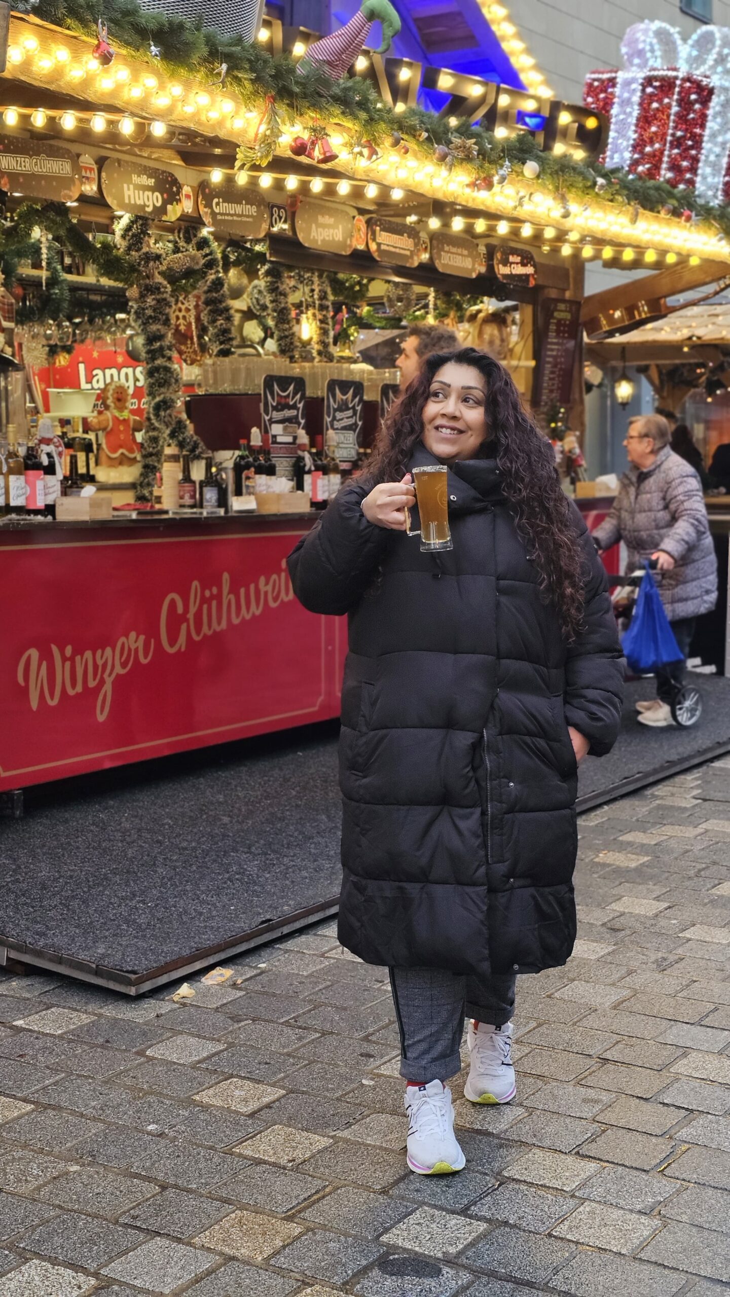 Nishi smiles whilst holding a glass mug of hot drink at a festive German Christmas market. She wears a long black puffer coat and white trainers, standing in front of a brightly lit mulled wine stall decorated with fir garlands and Christmas ornaments.