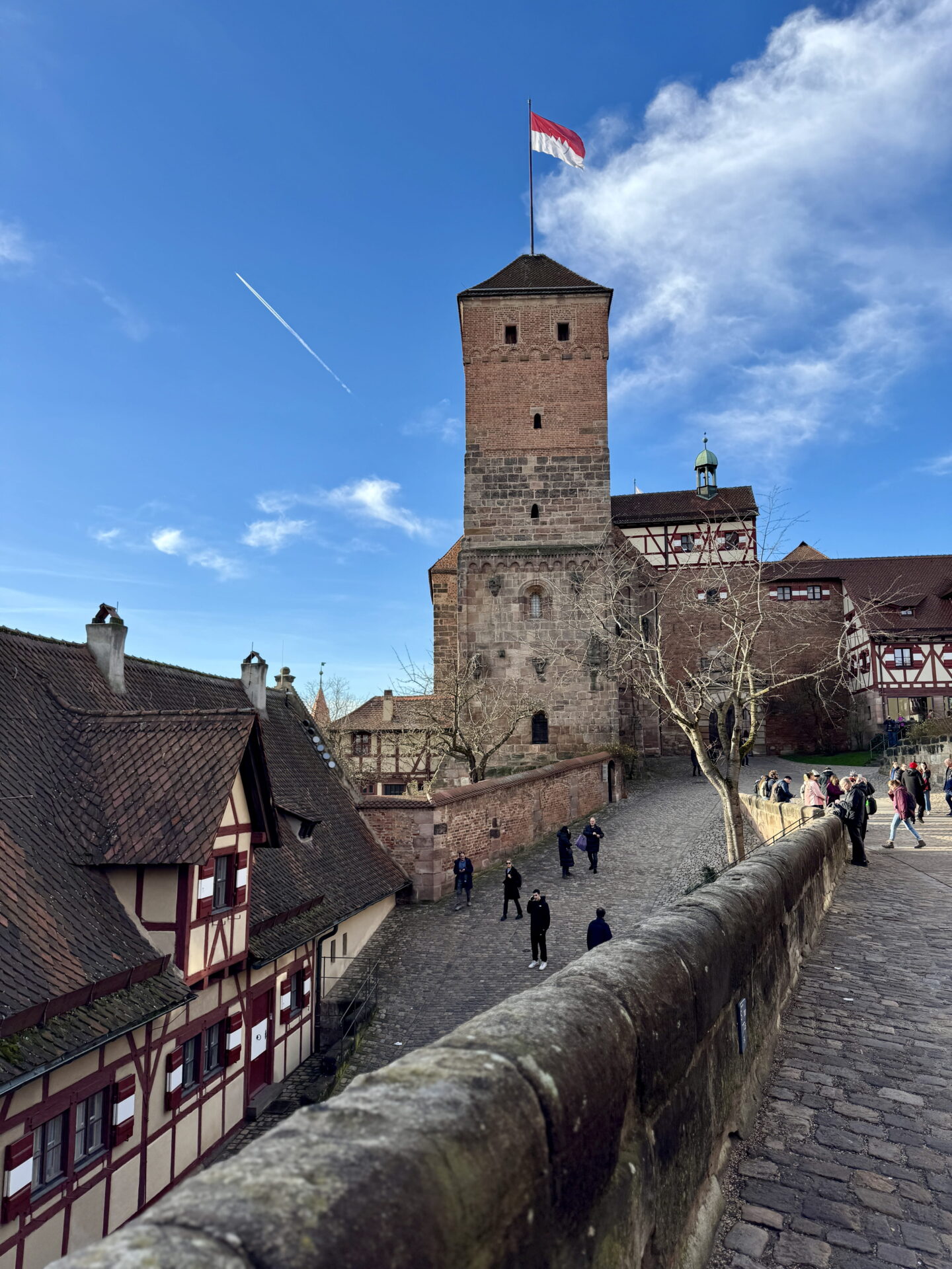 A view of Nuremberg Castle with its tall medieval stone tower flying a red and white flag above traditional half timbered buildings. Visitors explore the cobbled castle courtyard and raised stone walkway beneath a bright blue sky.