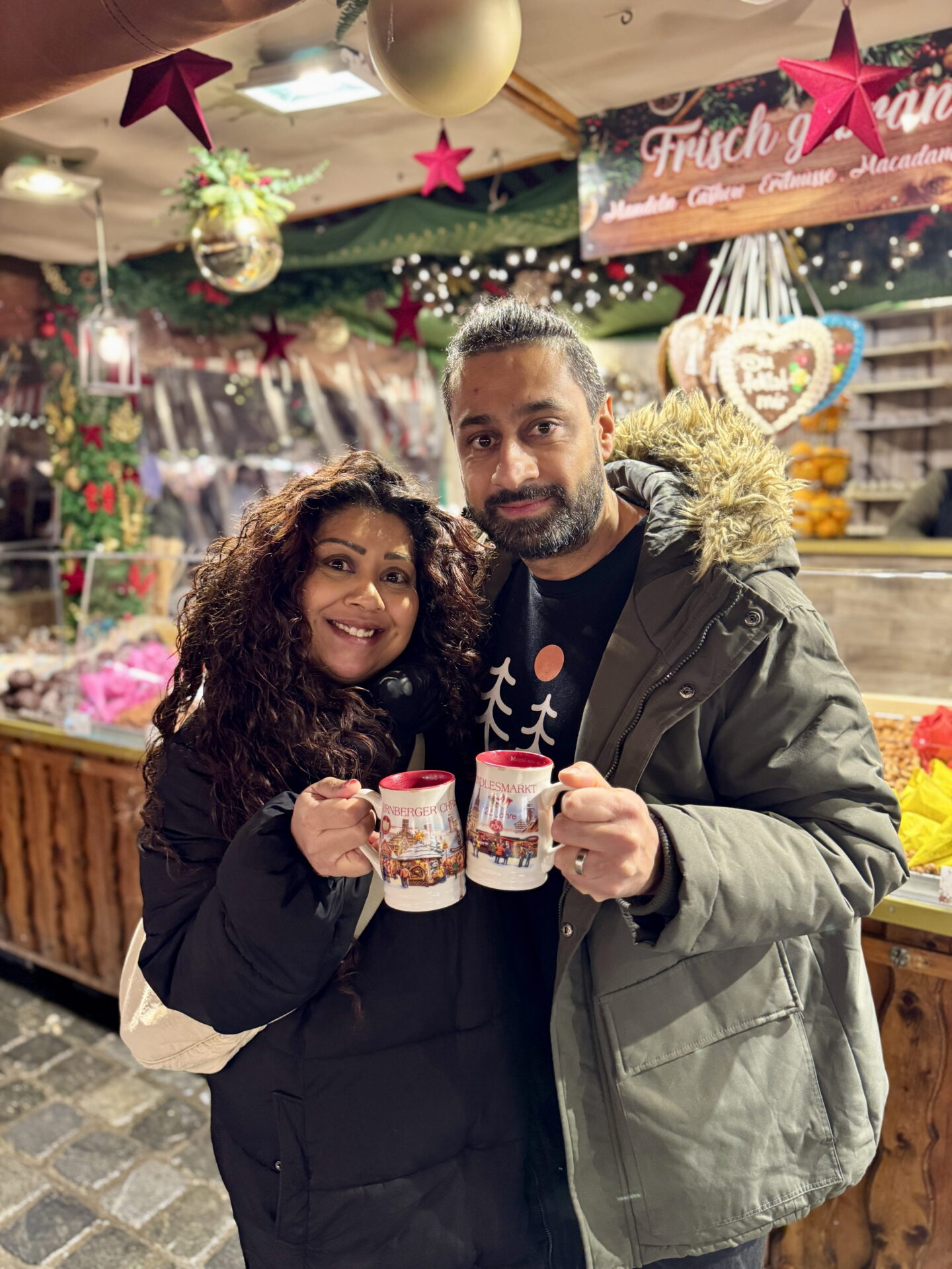 Nishi and Jass smile at the camera whilst holding illustrated Christmas market mugs in front of a brightly decorated festive sweets stall in Nuremberg. Nishi wears a black puffer coat and Jass wears a green parka with a fur trimmed hood, surrounded by hanging gingerbread hearts and Christmas decorations.