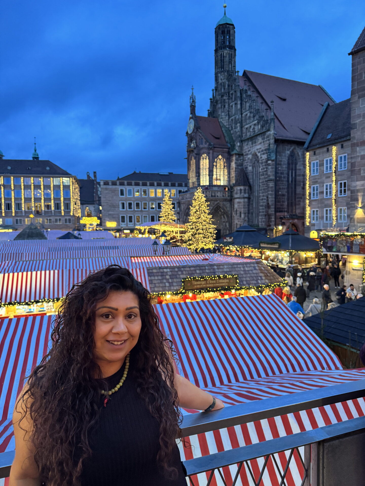 Nishi smiles whilst leaning against a balcony railing overlooking Nuremberg Christmas market at dusk. Rows of red and white striped market stalls, illuminated Christmas trees and the Gothic Frauenkirche fill the historic square behind her.