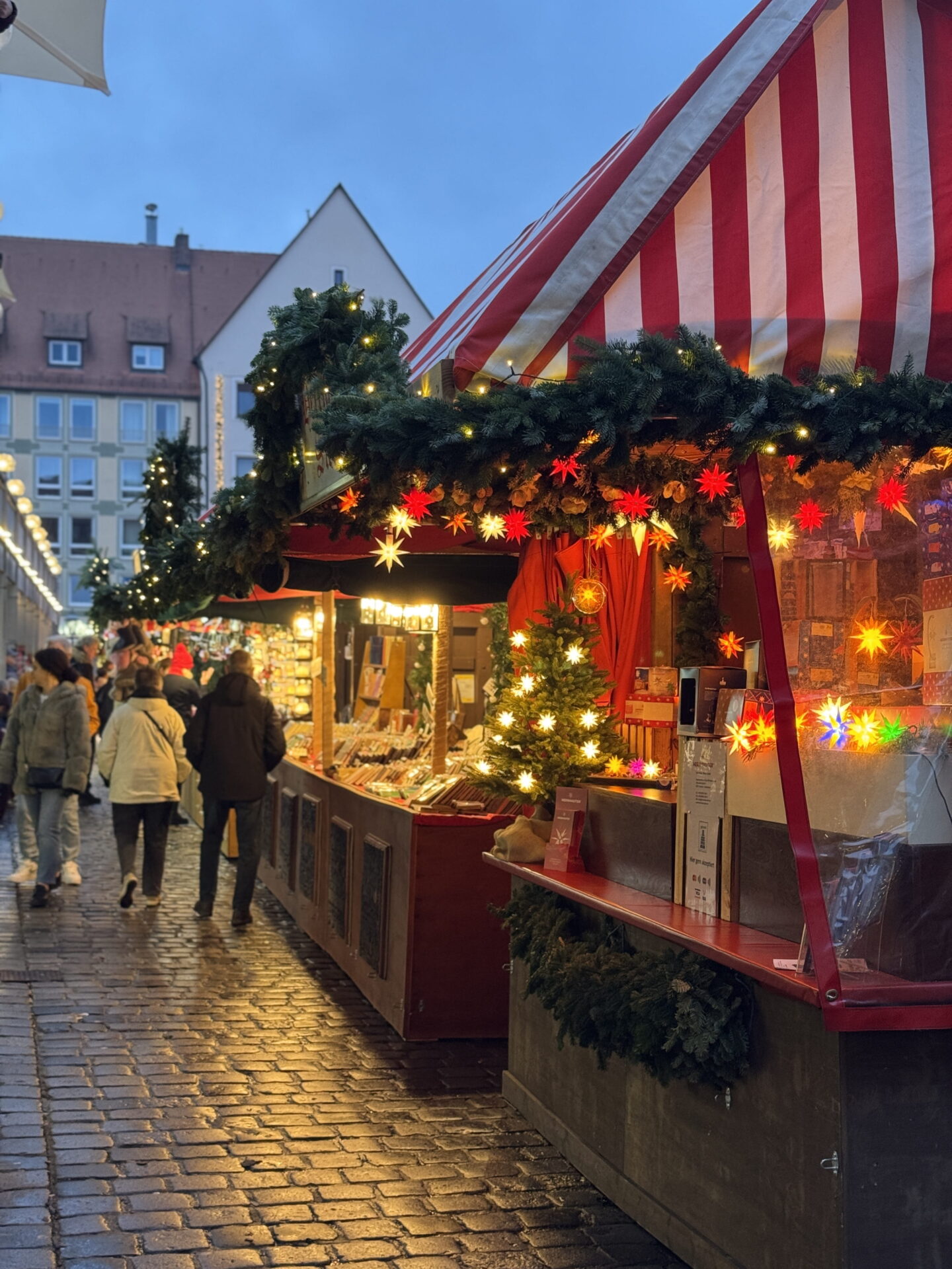 A row of wooden Christmas market stalls with red and white striped roofs glows with warm fairy lights and colourful star decorations. Shoppers stroll along the wet cobbled street beneath fir garlands and festive displays as evening falls.