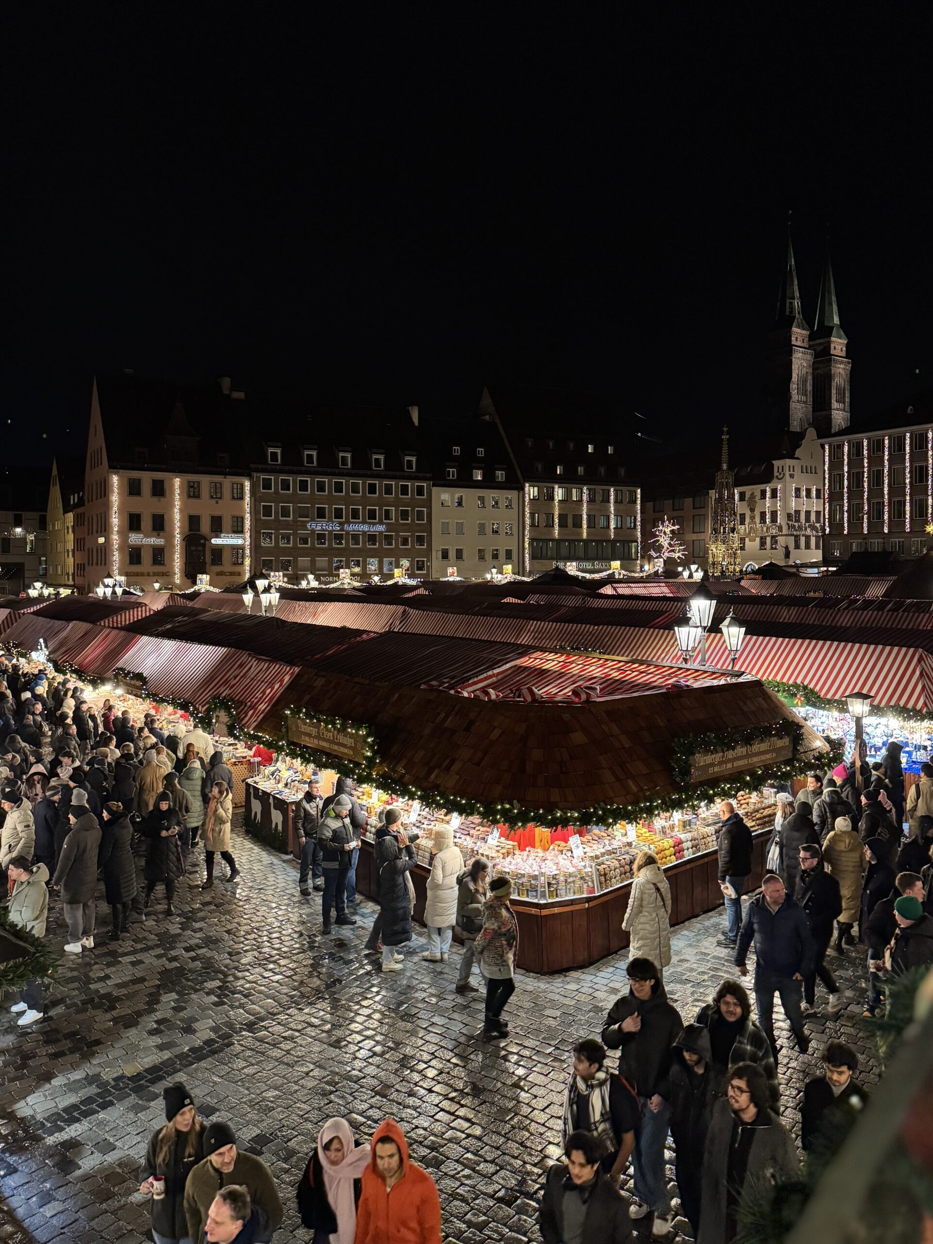 An elevated view of Nuremberg Christmas market at night, with crowds browsing brightly illuminated wooden stalls beneath red and white striped roofs. Historic buildings and the twin towers of St Sebaldus Church rise above the festive square against the dark sky.