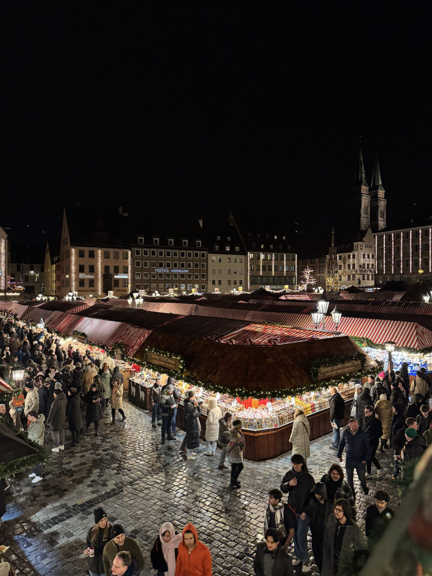 An elevated view of Nuremberg Christmas market at night, with crowds browsing brightly illuminated wooden stalls beneath red and white striped roofs. Historic buildings and the twin towers of St Sebaldus Church rise above the festive square against the dark sky.