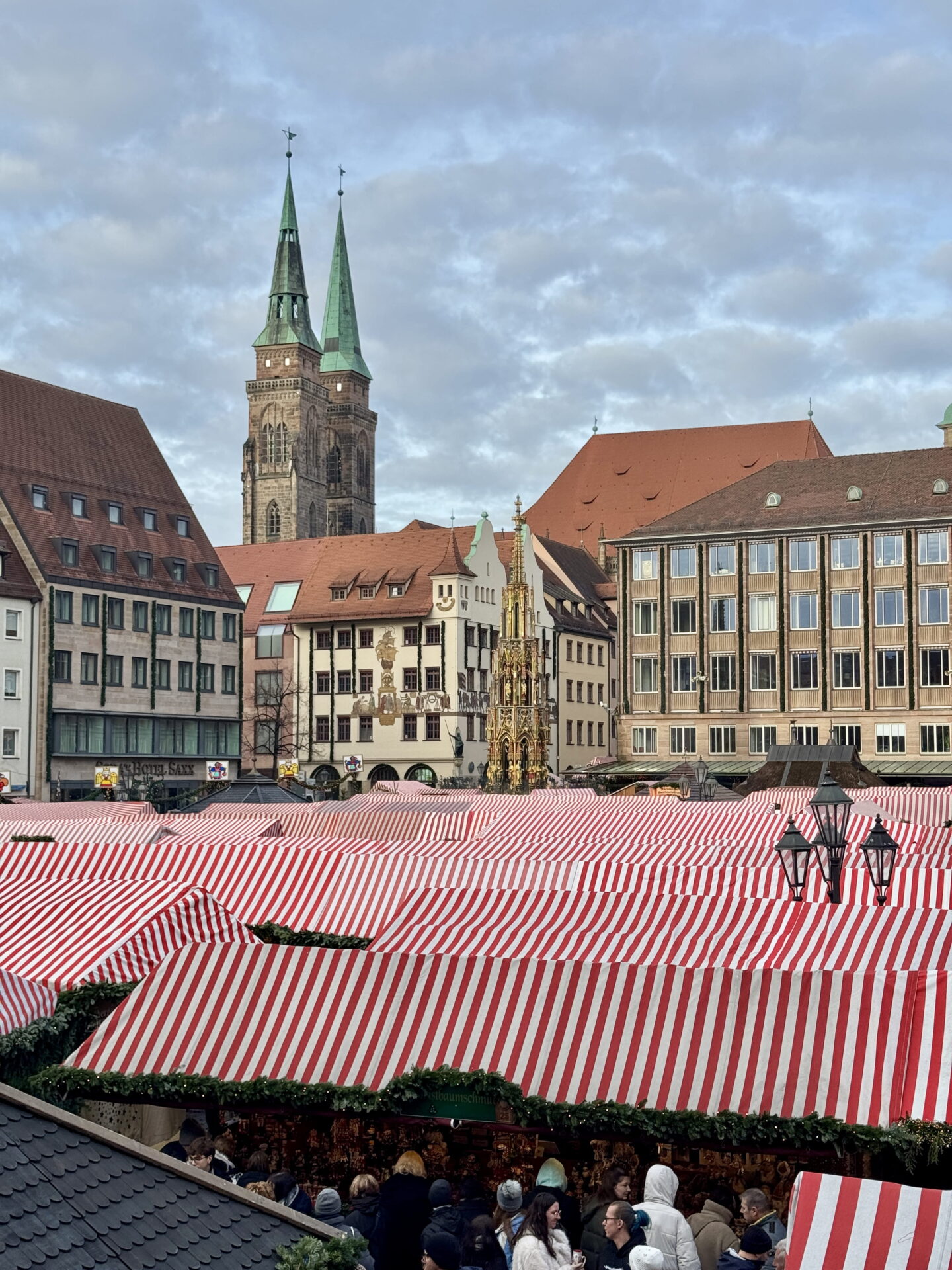 Rows of red and white striped Christmas market stalls fill Nuremberg main square beneath a cloudy sky. The twin spires of St Sebaldus Church and the ornate golden Schöner Brunnen fountain stand among the historic buildings overlooking the market.