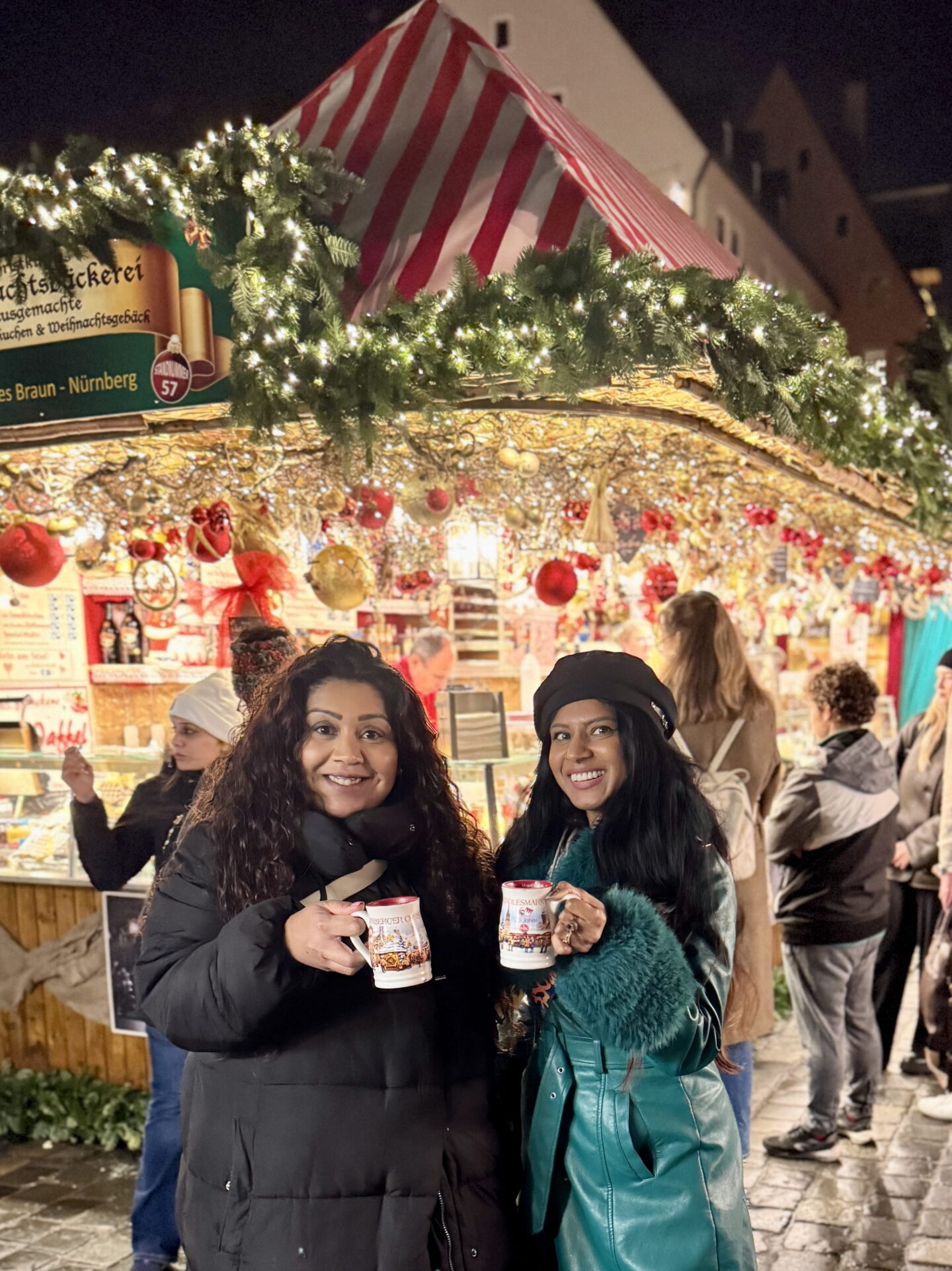 Nishi and Priya smile at the camera whilst holding illustrated Christmas market mugs in front of a brightly lit festive stall at night. Nishi wears a black puffer coat whilst Priya wears a green coat with fluffy cuffs and a black beret, surrounded by hanging red baubles and Christmas decorations.