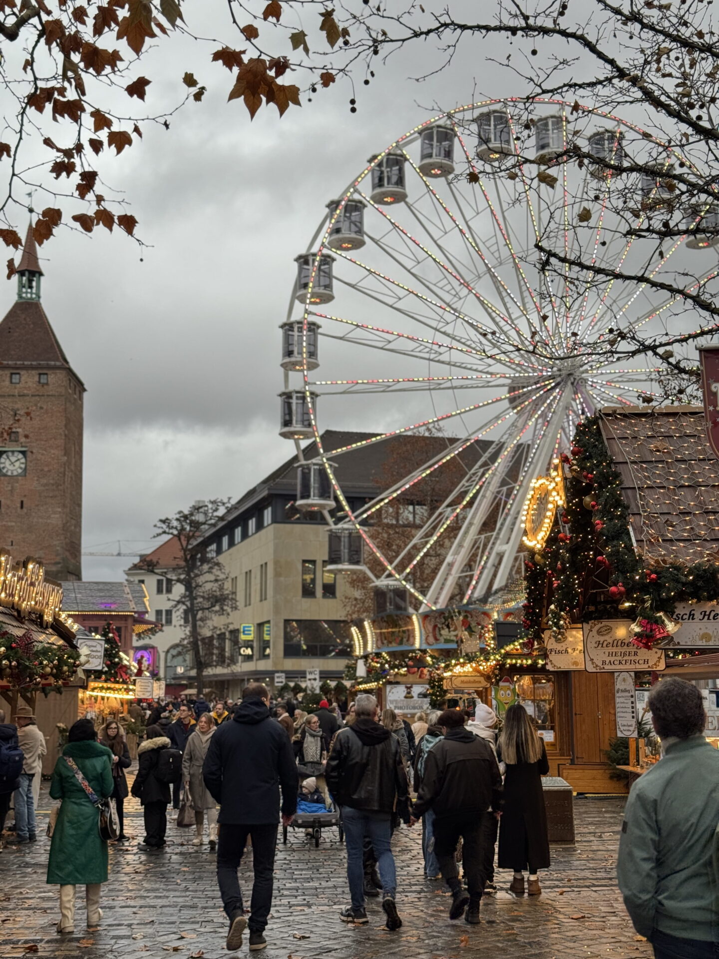 A large illuminated Ferris wheel towers above a busy German Christmas market, with visitors walking between wooden stalls on a wet cobbled street. Bare tree branches frame the scene, with a historic brick clock tower standing to the left beneath an overcast sky.
