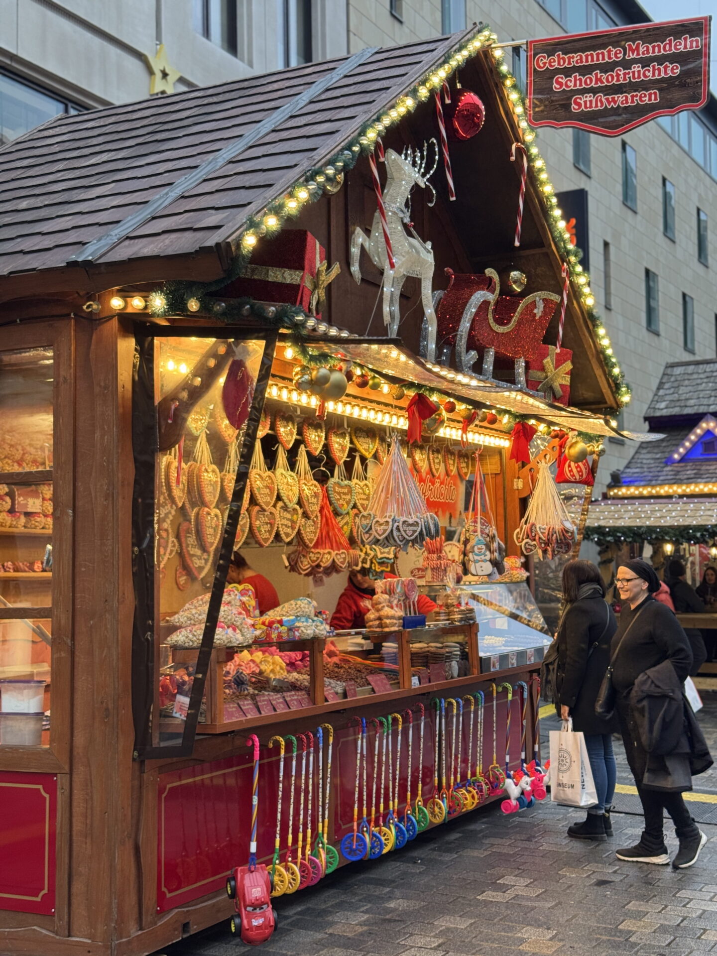 A brightly illuminated wooden Christmas market stall displays colourful sweets, candy canes and hanging gingerbread hearts beneath festive reindeer decorations and fir garlands. A sign above the stall reads "Gebrannte Mandeln Schokofrüchte Süßwaren" as shoppers browse the treats.