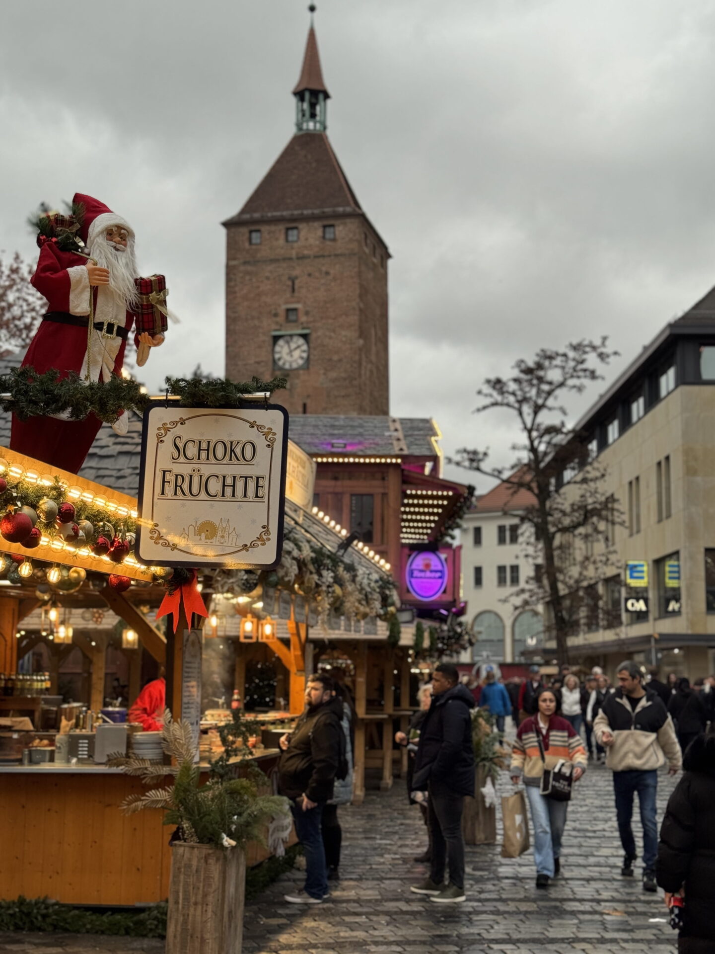 A Father Christmas figure dressed in red stands above a wooden Christmas market stall decorated with fairy lights, fir garlands and baubles. A hanging sign reads "Schoko Früchte" with a historic brick clock tower and shoppers visible along the cobbled street behind it.