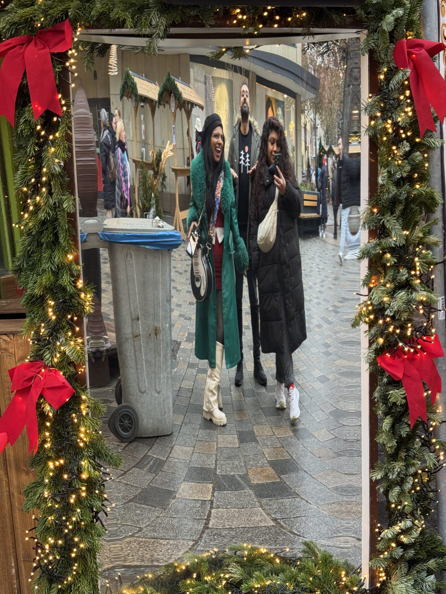 Priya, Jass and Nishi pose for a mirror selfie at a Christmas market, framed by fir garlands, glowing fairy lights and large red bows. Priya wears a green coat and white knee high boots, Jass stands between them in a dark coat and Nishi holds her phone whilst wearing a black puffer coat.