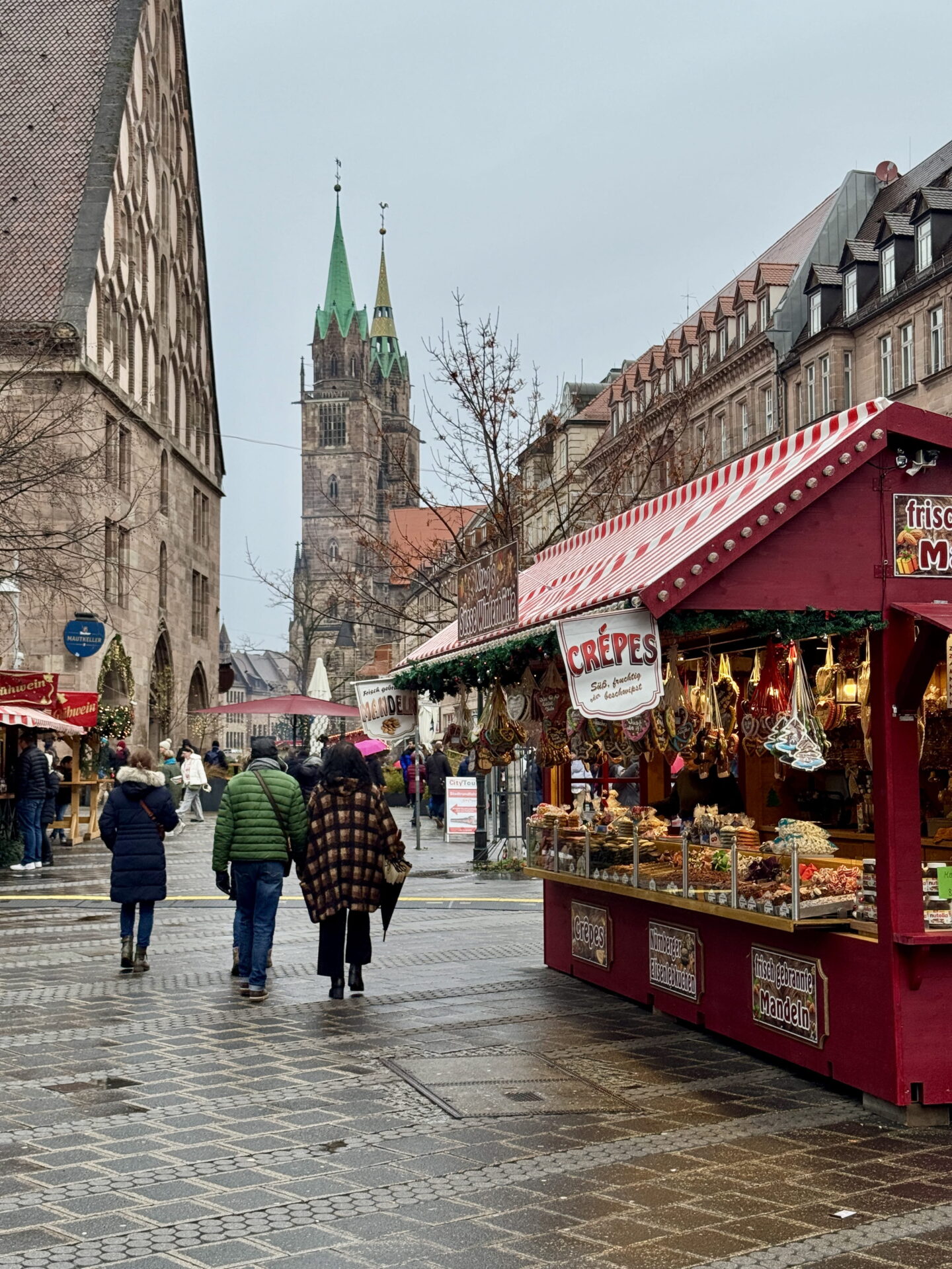 Visitors walk along a wet cobbled street at a Christmas market in Nuremberg, passing red wooden stalls selling crepes, roasted almonds and festive treats. The twin green spires of St Sebaldus Church rise above the historic buildings in the background.