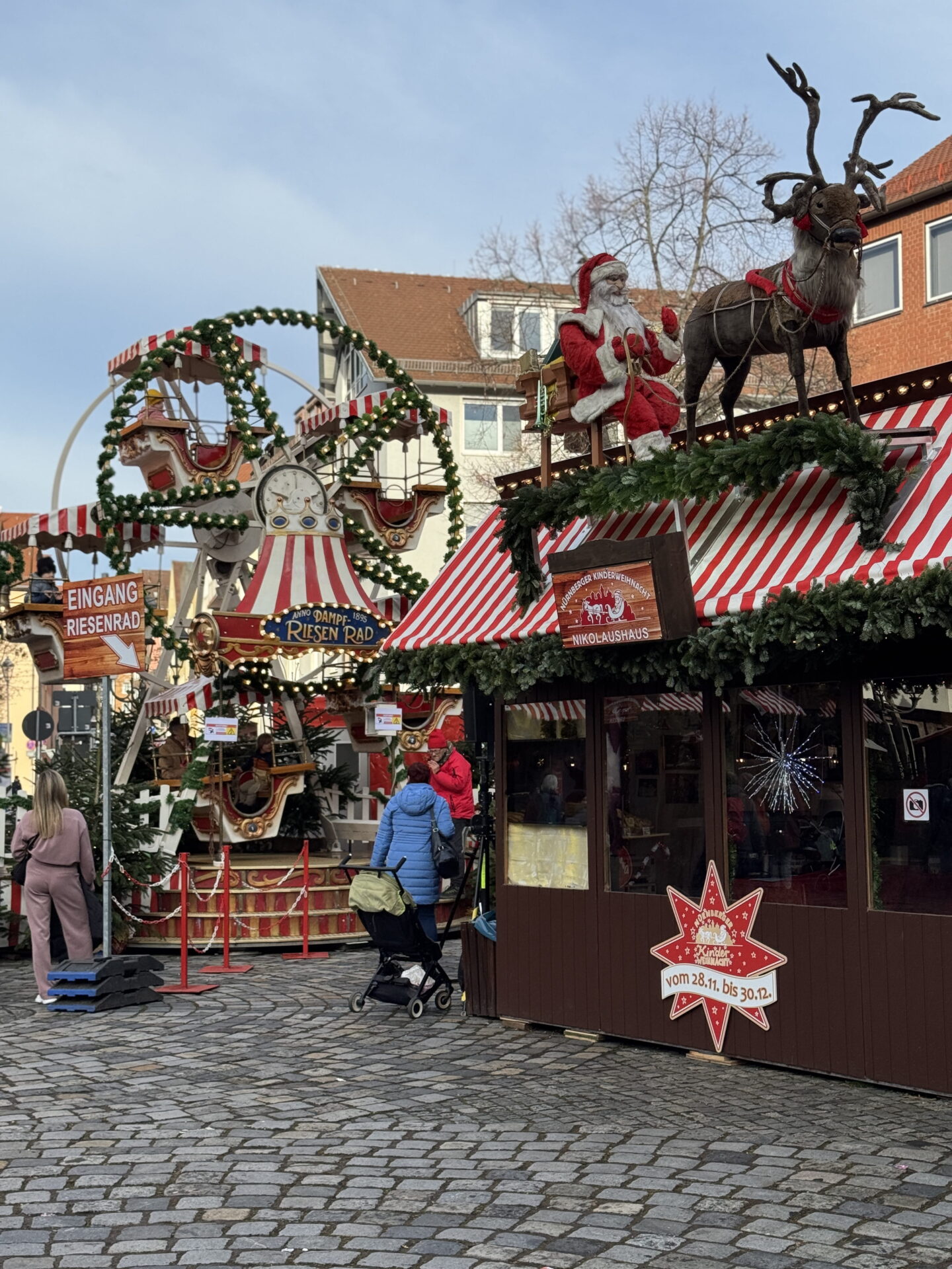 A traditional children's Ferris wheel decorated with red and white striped canopies and green Christmas garlands stands beside a wooden market hut topped with Father Christmas and a reindeer. The festive attraction at Nuremberg Christmas market has signs reading "Eingang Riesenrad" and "Nikolaushaus".
