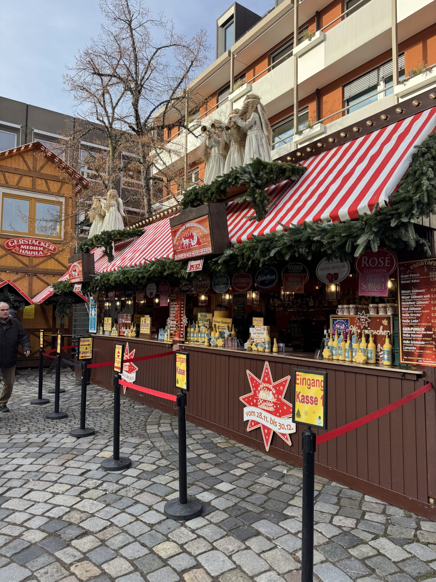 A row of wooden Christmas market drinks stalls with red and white striped roofs, fir garlands and white angel decorations above the counters. Signs advertise mulled wine, hot chocolate and other festive drinks, with red queue barriers arranged along the cobbled street.