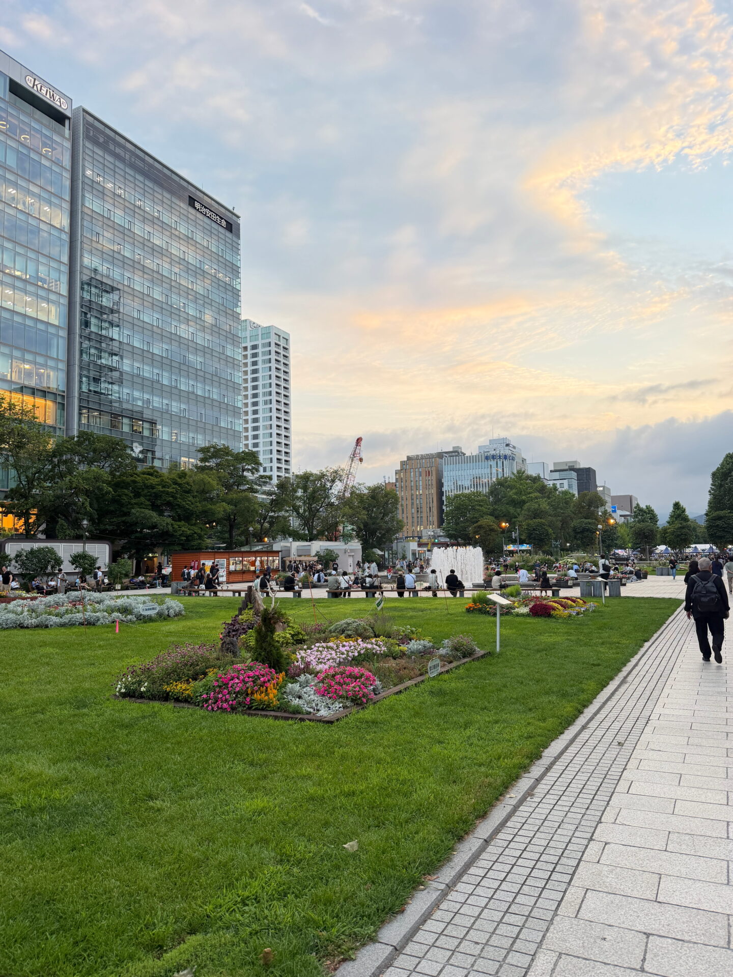 Odori Park in central Sapporo at sunset with neatly maintained lawns, colourful flowerbeds and a large fountain surrounded by visitors. Modern office buildings rise behind the trees beneath a soft pink and golden evening sky.