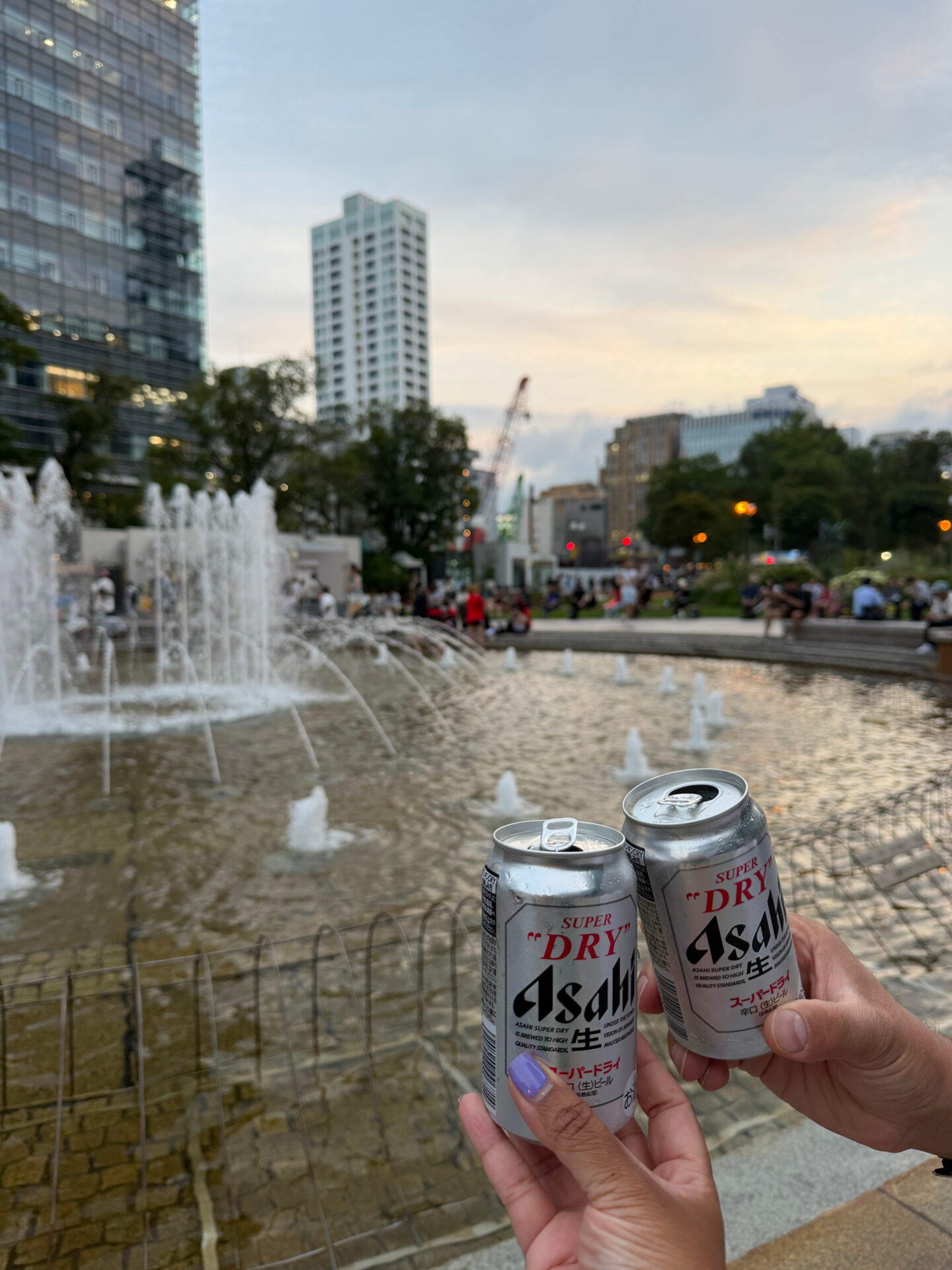 Two people clink silver cans of Asahi Super Dry beer beside a large fountain in Odori Park in Sapporo. Water jets spray into the pool while visitors relax around the park in the early evening.